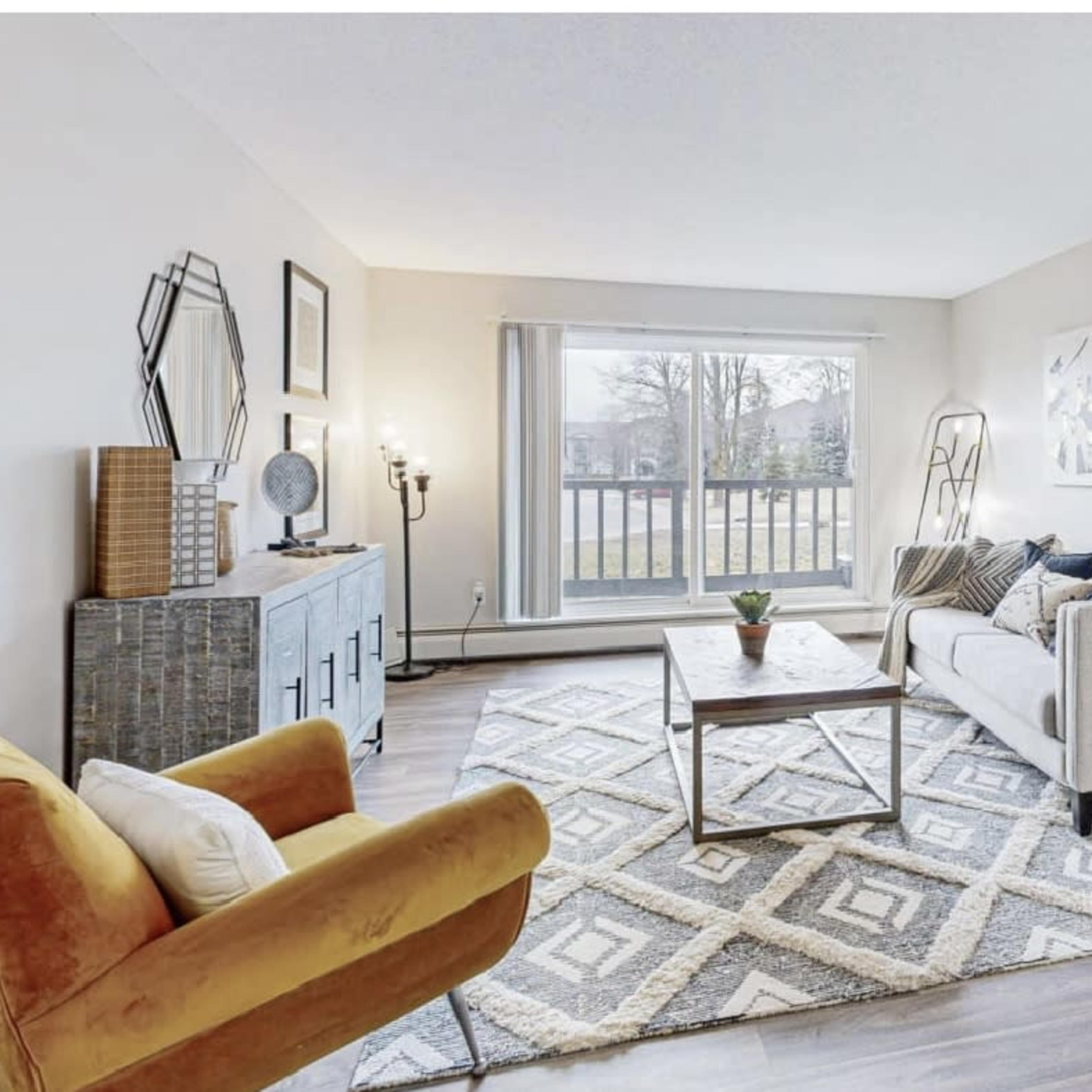 A modern living room features a light-colored sofa, an orange armchair, a coffee table, and a large window with a view of the outdoors.