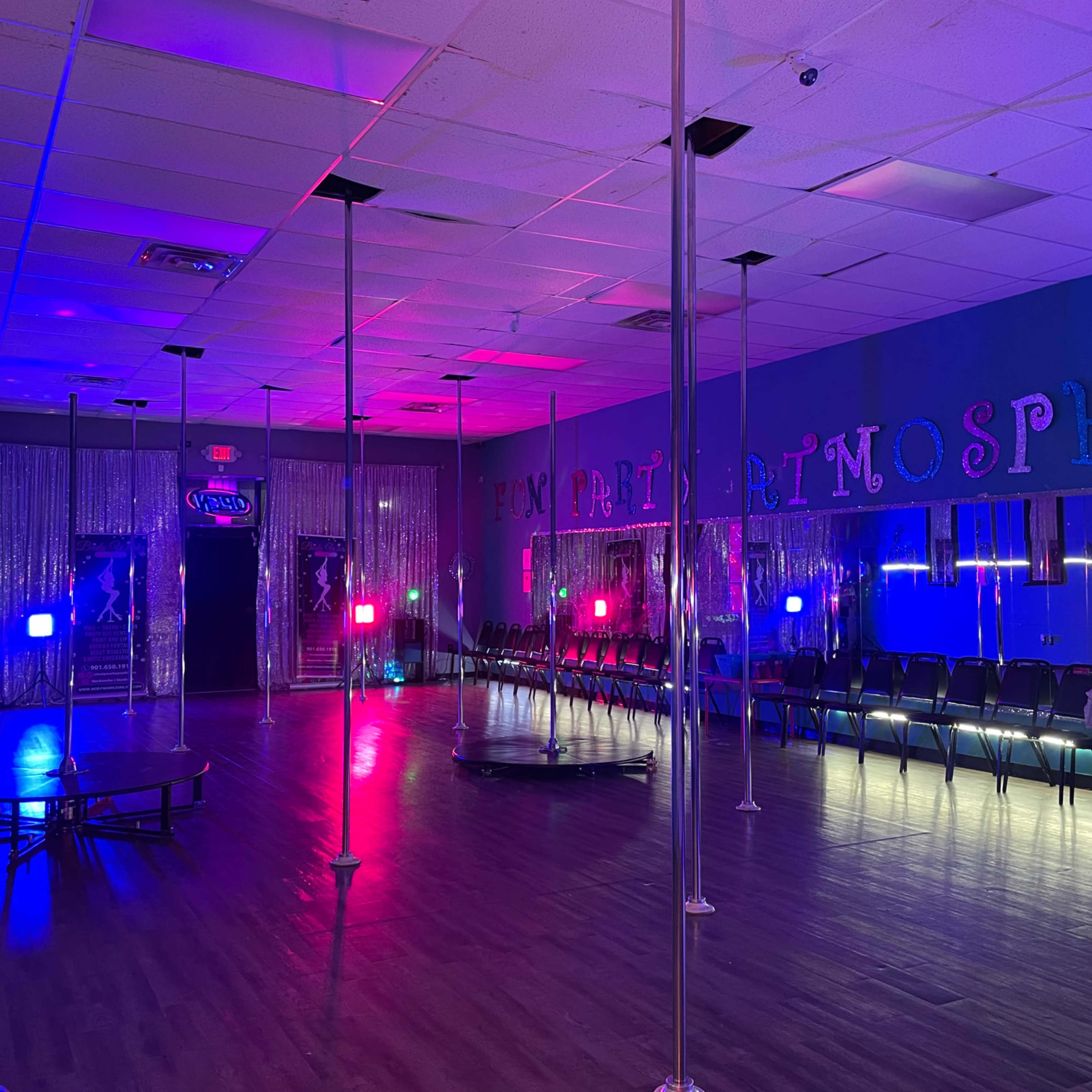 A brightly lit dance studio featuring several poles and rows of chairs along the sides, with colorful lights illuminating the space.