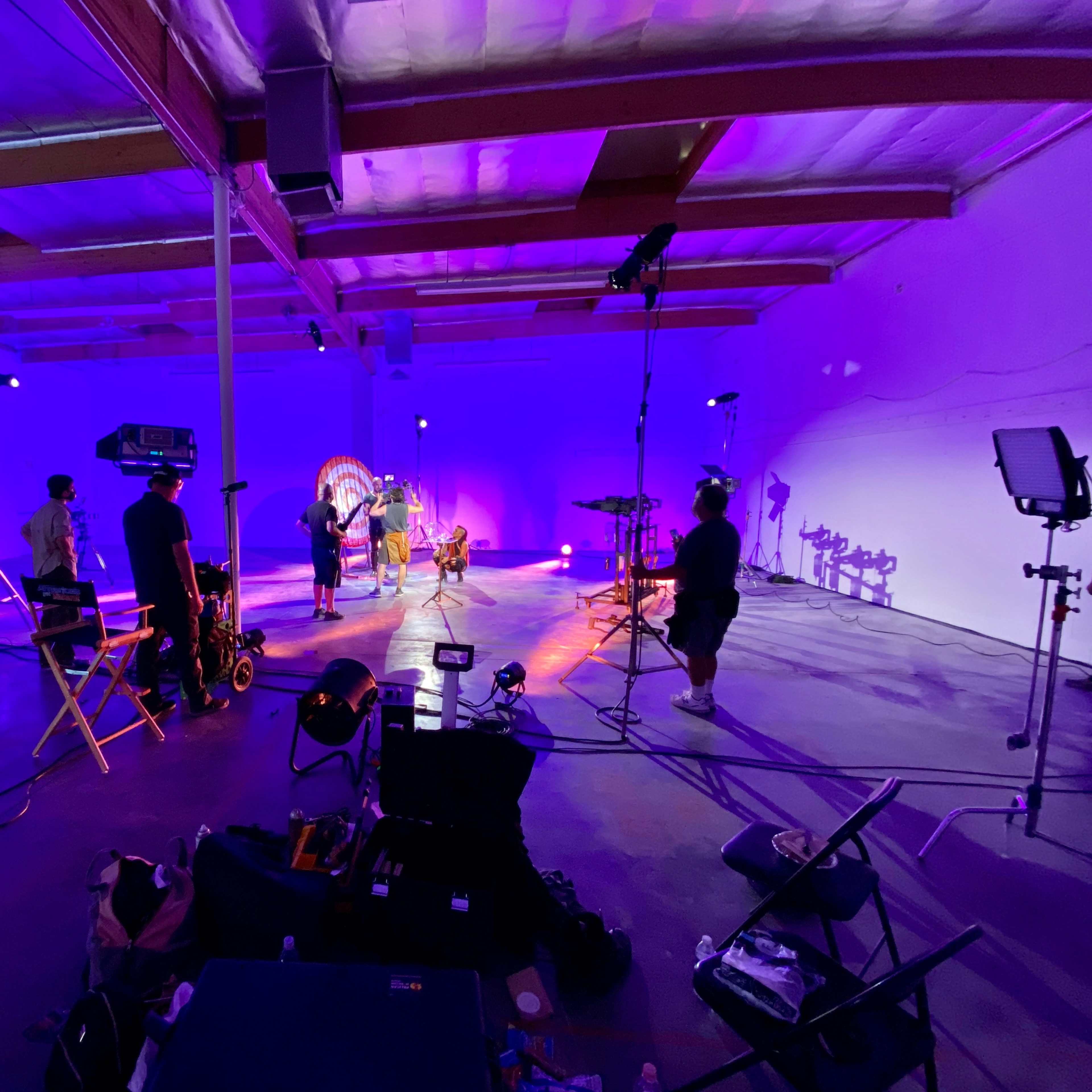 A group of people is setting up equipment and performing in a brightly lit, purple-toned studio space.