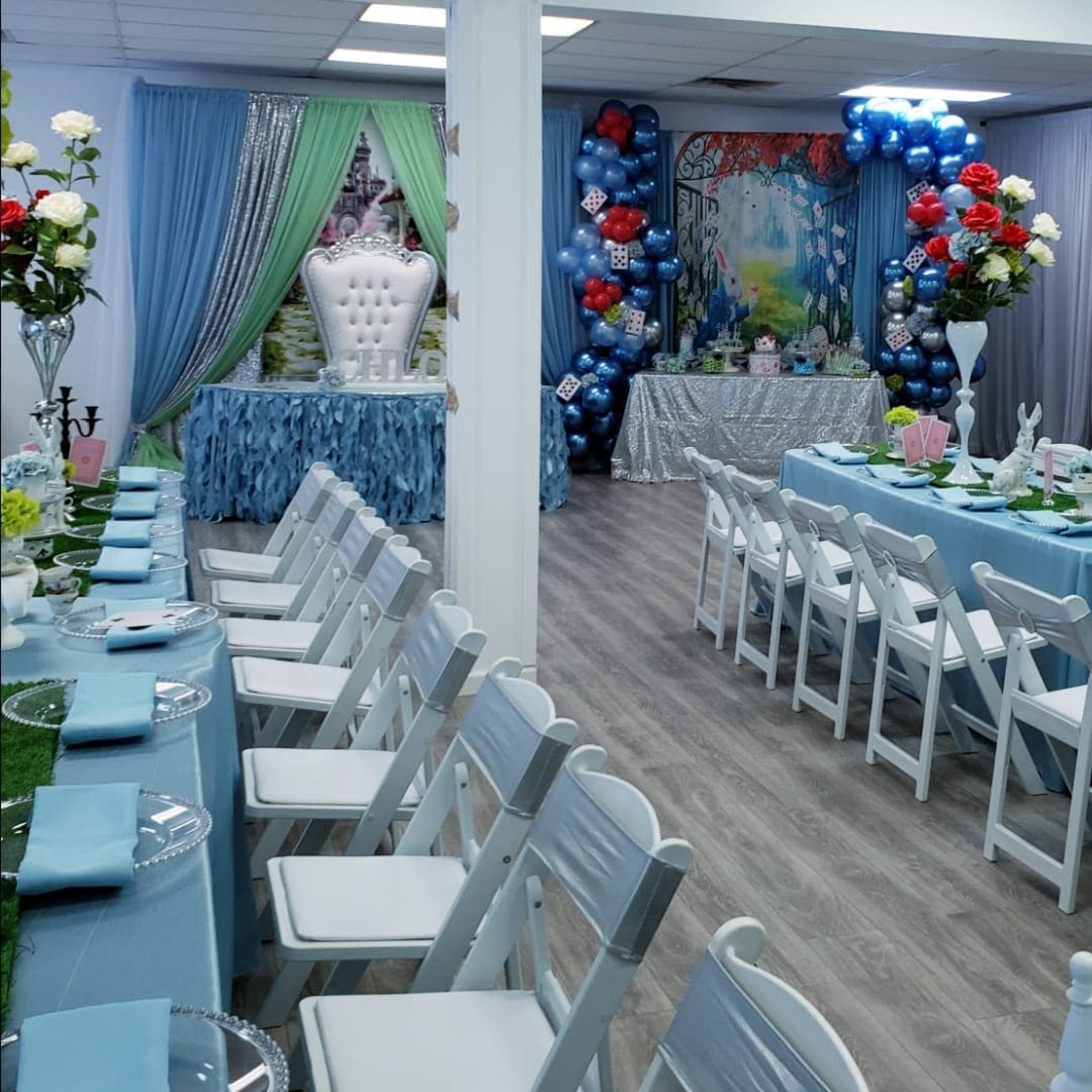 A decorated event space with tables set for a celebration, featuring white chairs, blue tablecloths, and colorful balloon arrangements.