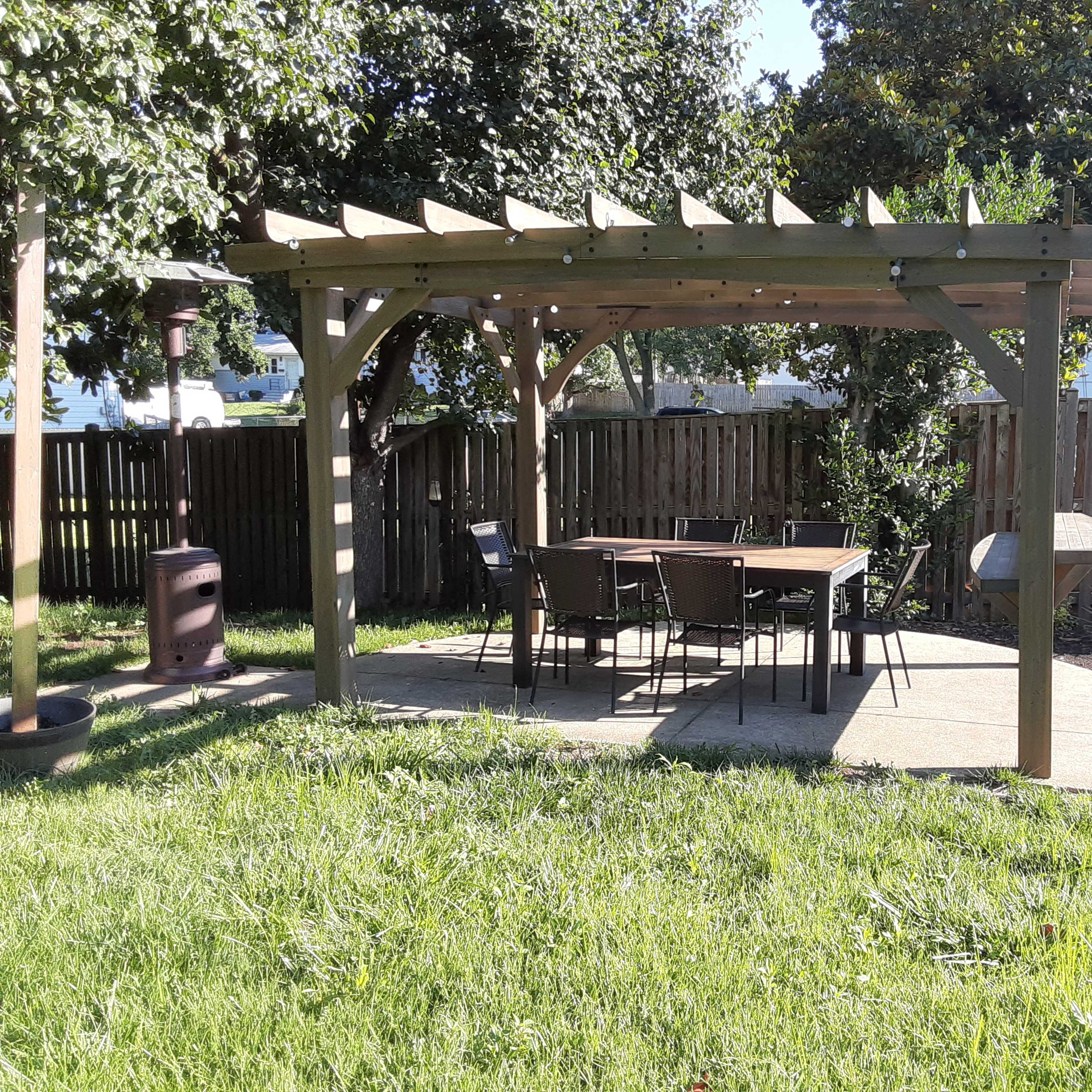 A wooden pergola with a table and chairs is set on a concrete patio in a grassy backyard.