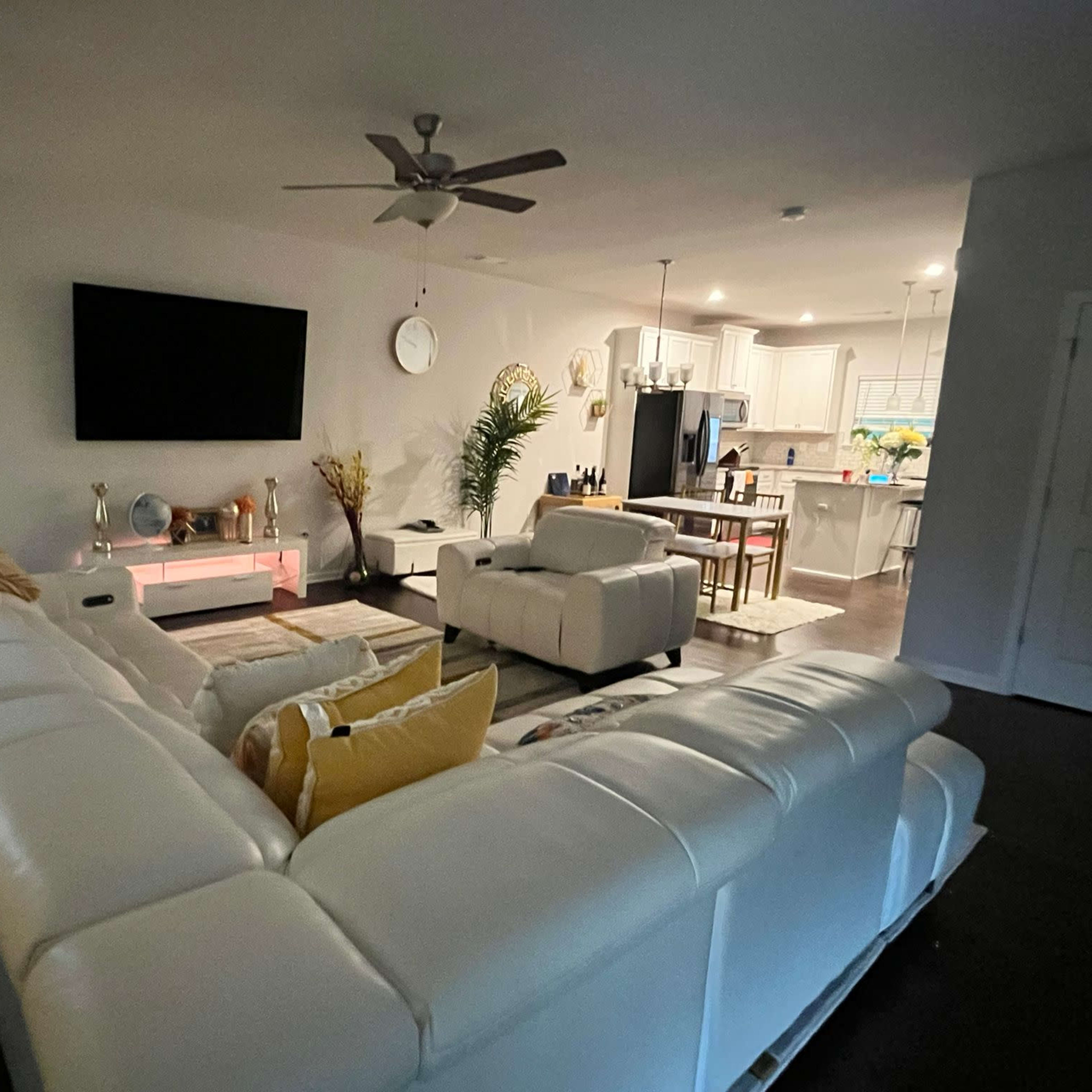 A modern living room with a white sectional sofa, a large wall-mounted television, and an open kitchen in the background.