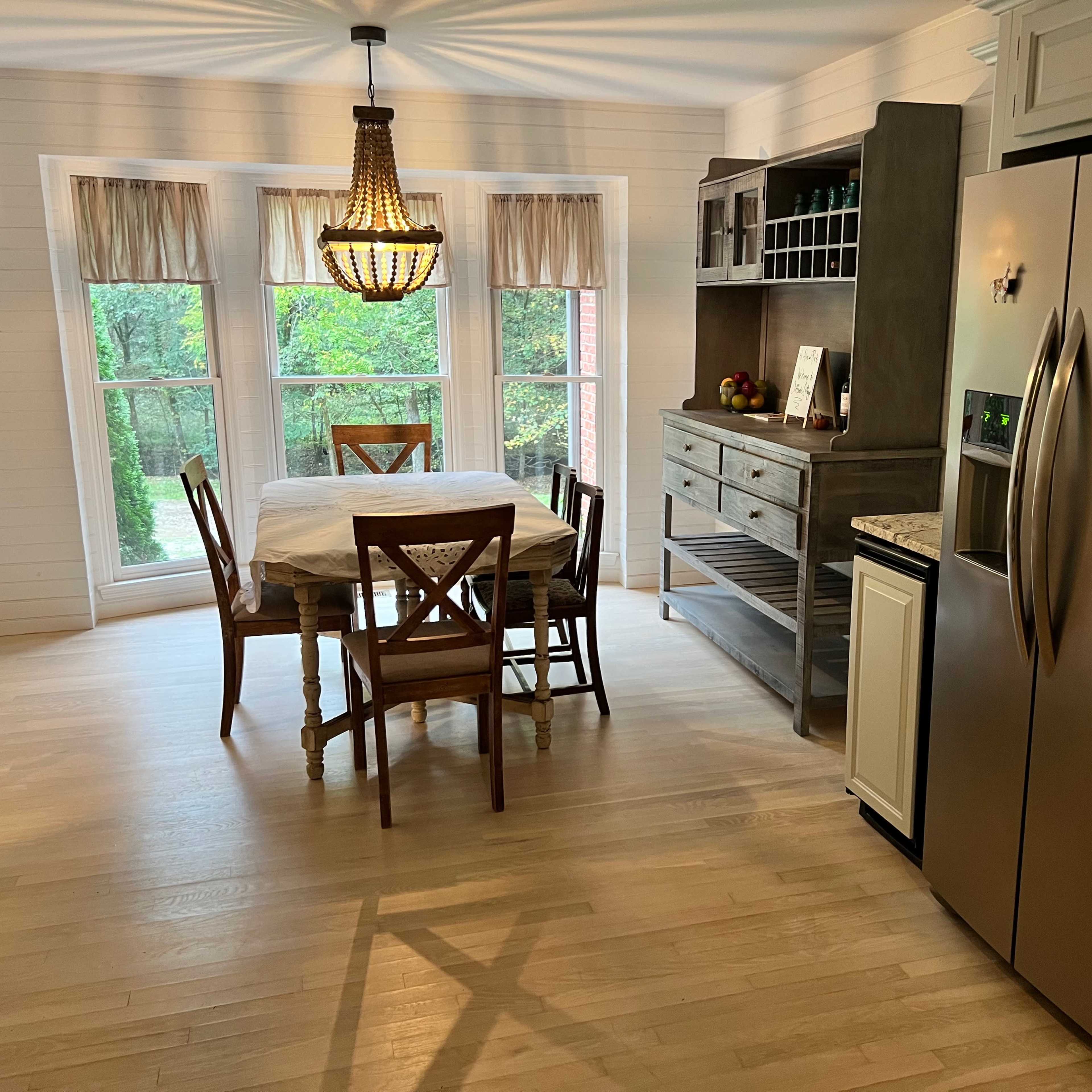A dining area features a wooden table with four chairs, a sideboard, and large windows overlooking a green landscape.