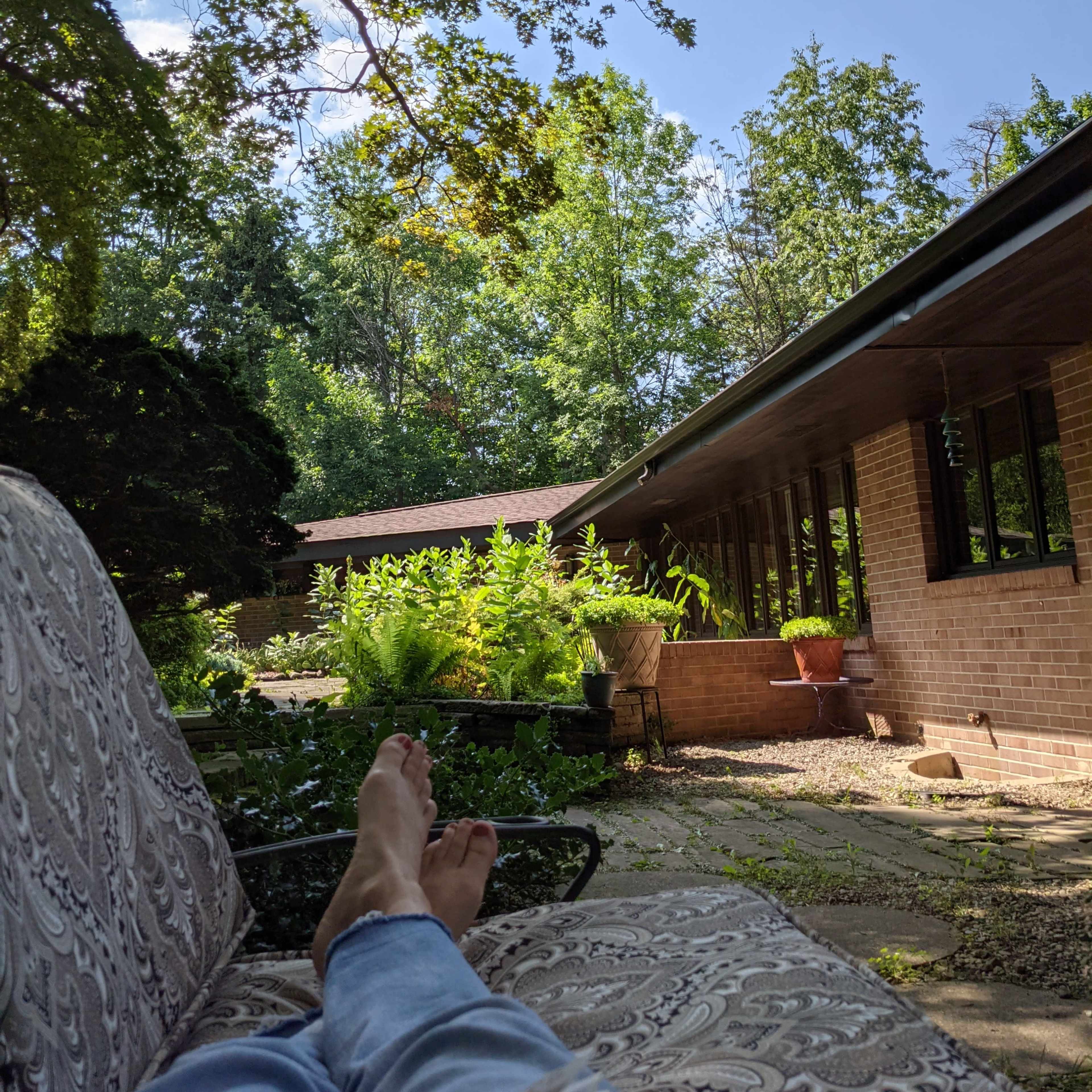 A pair of bare feet rests on a cushioned outdoor chair in a garden filled with lush greenery, with a brick house visible in the background.