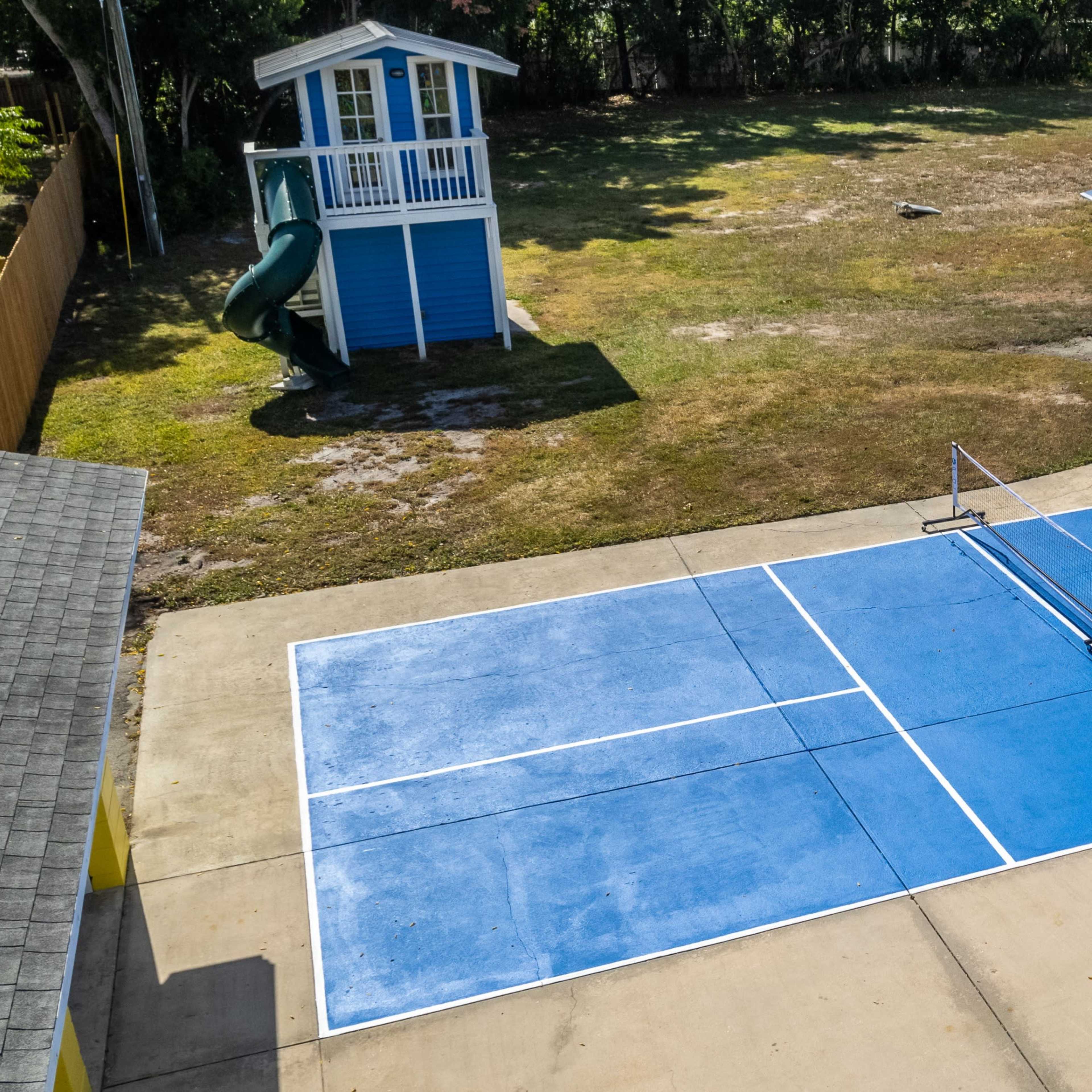 A blue outdoor tennis court adjacent to a house, with a play structure in the background.