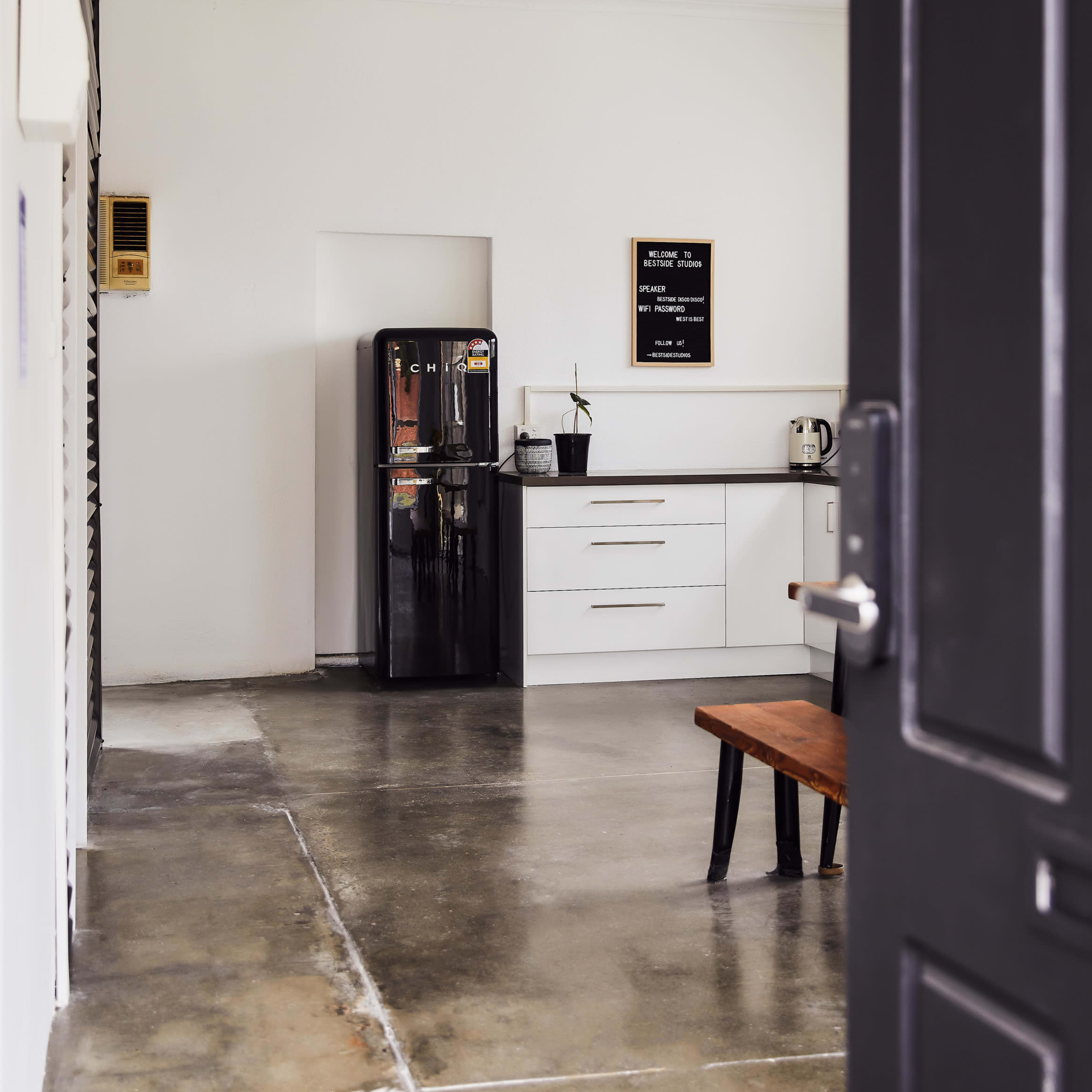 A modern kitchen space features a black refrigerator, a white countertop, and a wooden bench with polished concrete flooring.