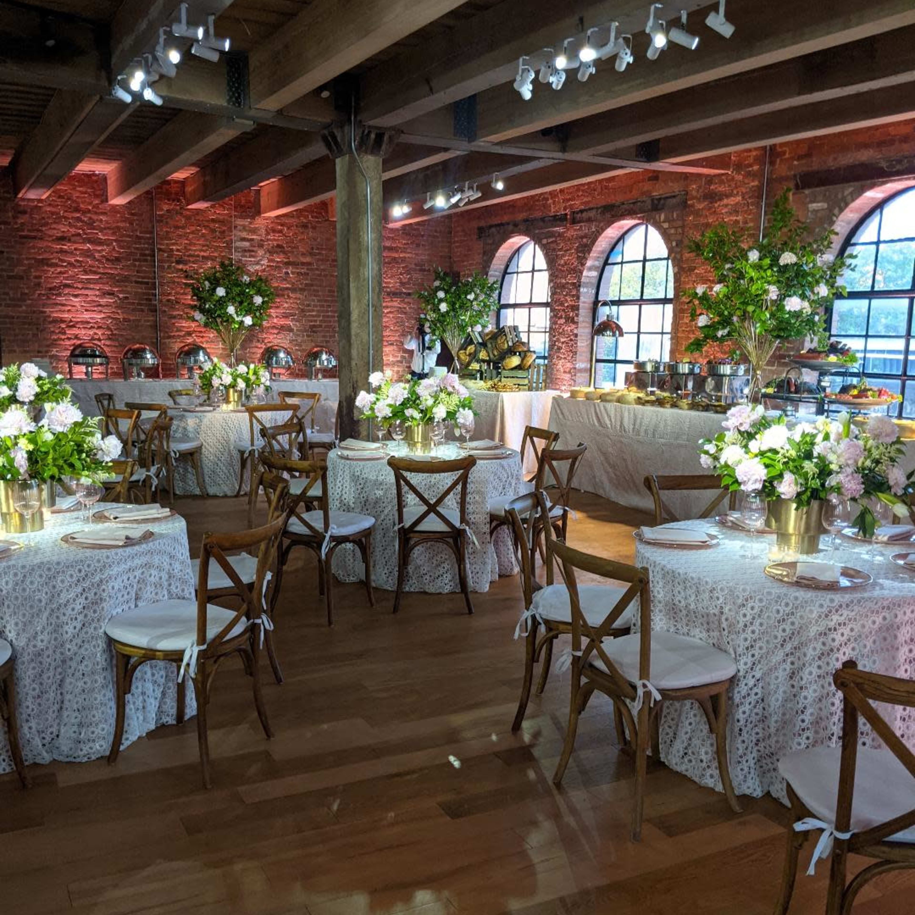 An elegantly arranged dining area with round tables covered in white cloth, decorated with flower centerpieces and surrounded by wooden chairs, all under soft lighting in a rustic venue.