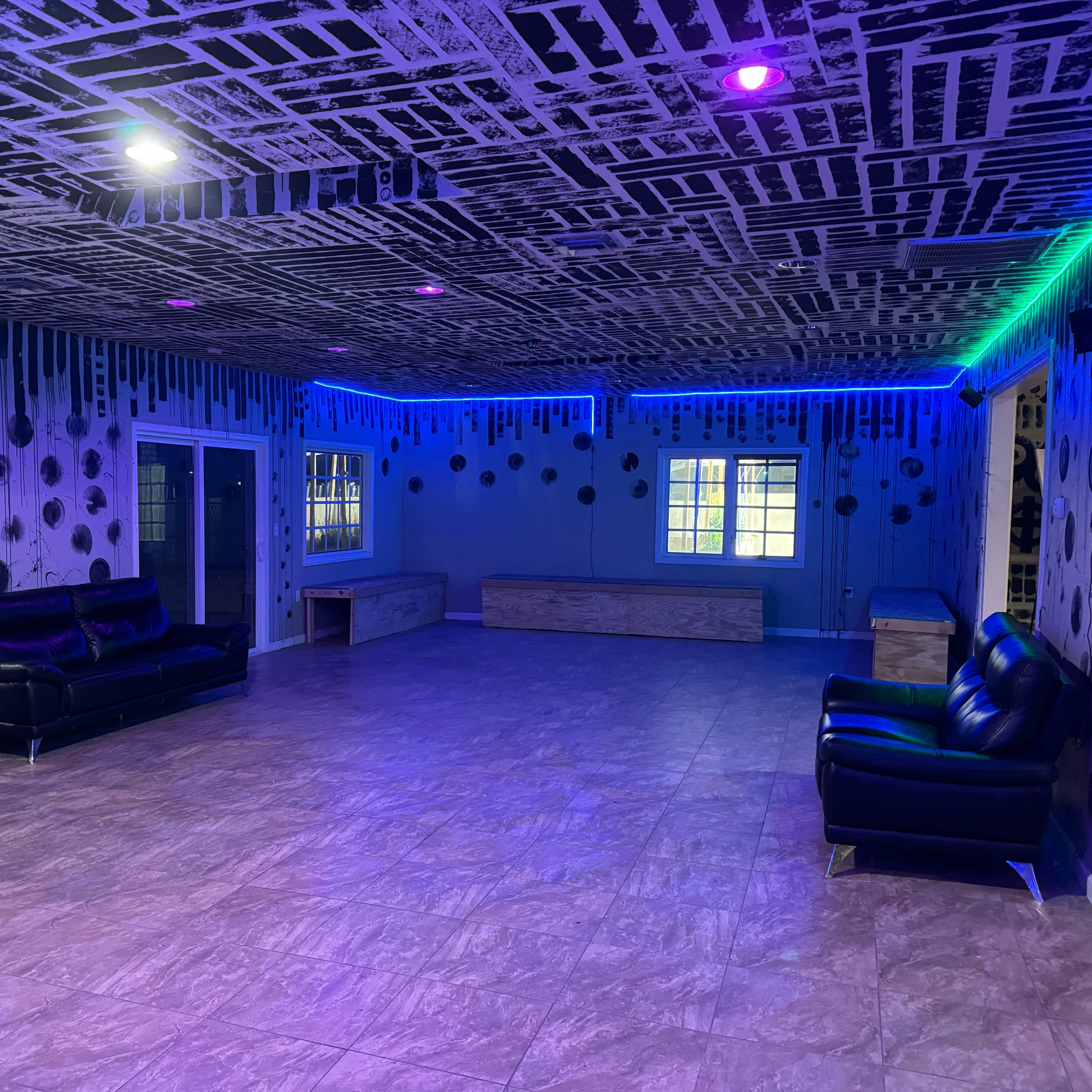 A spacious room with a patterned black and white ceiling, purple and green ambient lighting, and a few pieces of furniture, including sofas and tables.