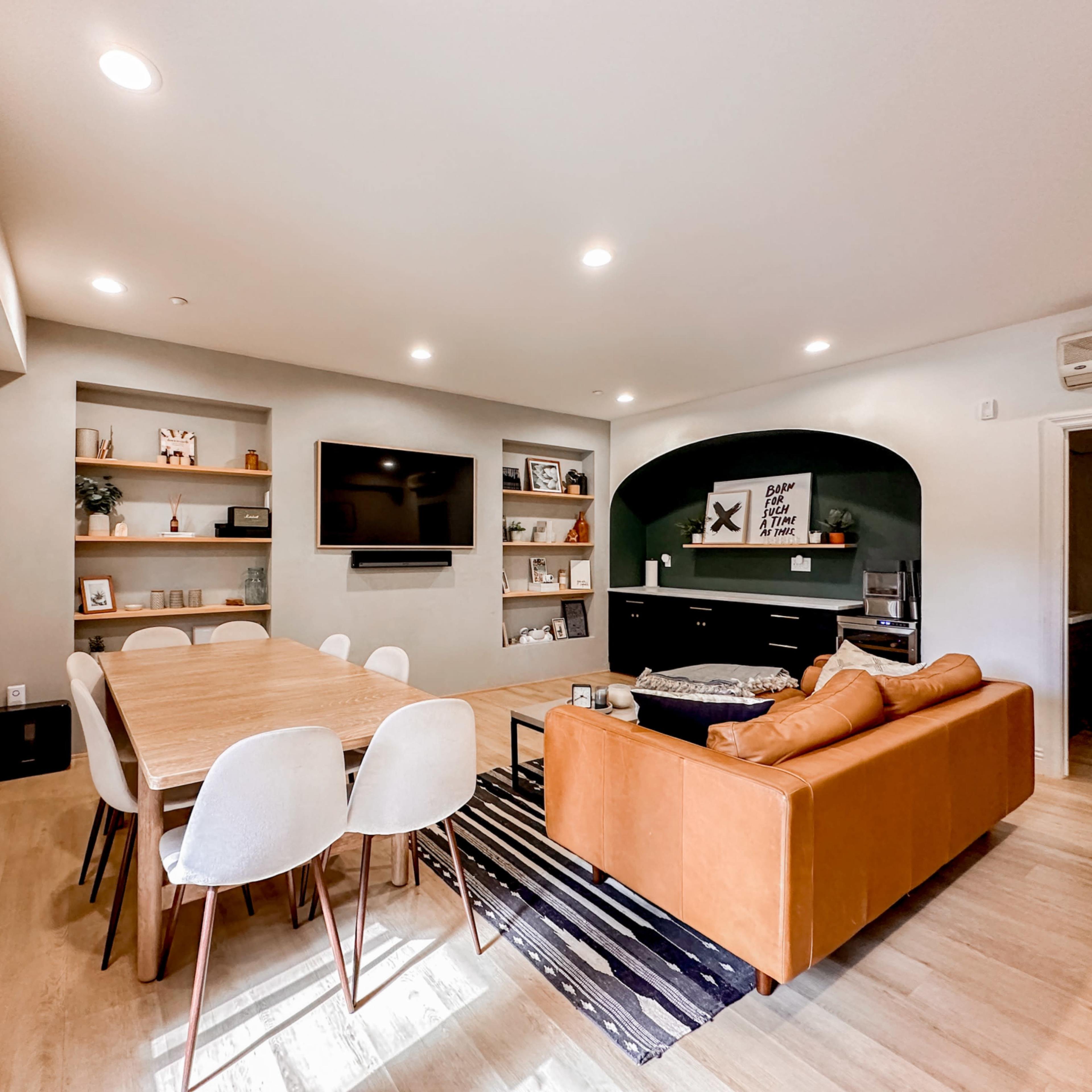 A modern living space with a dining table, a cozy sofa, shelves displaying decor, and a media center with a television.