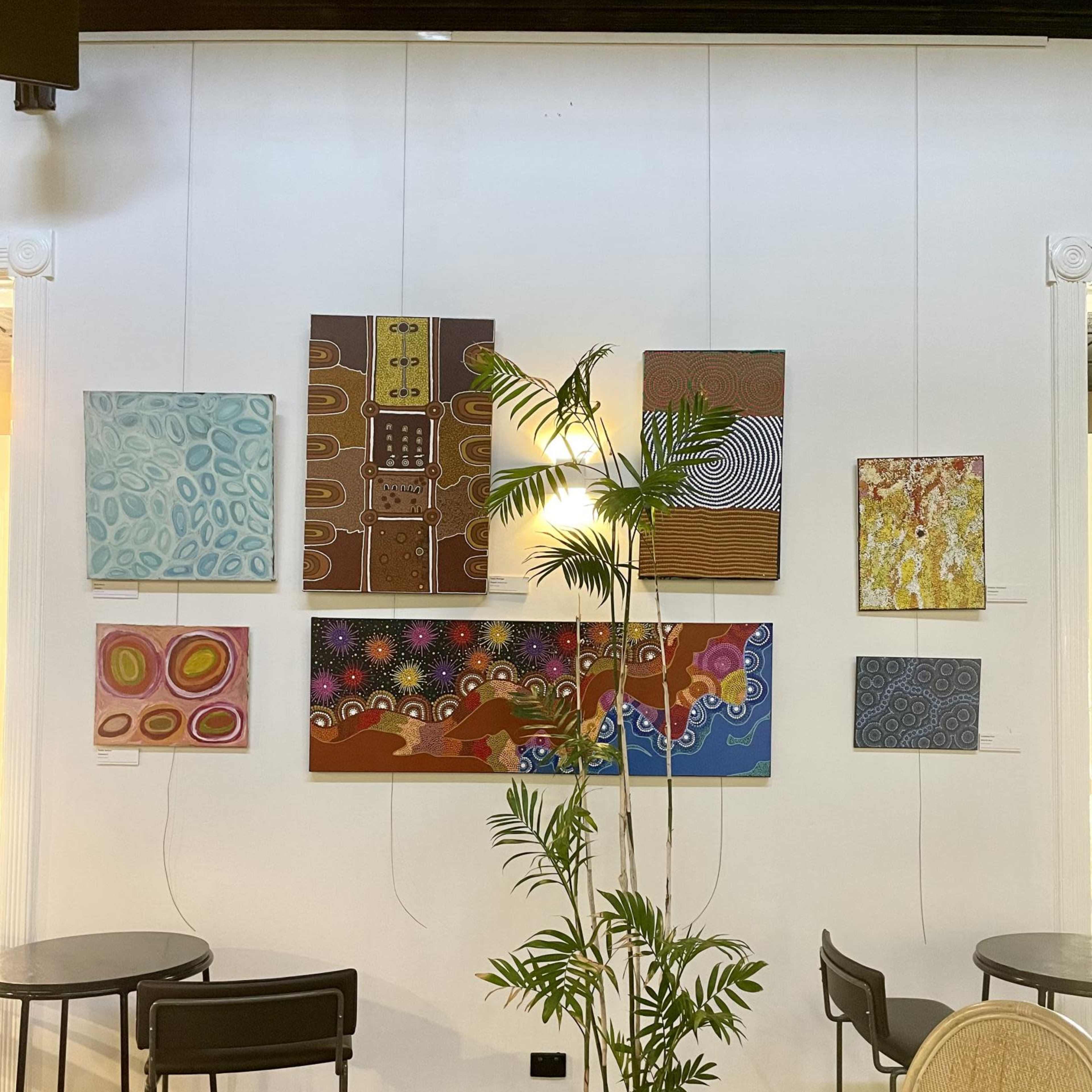 A gallery wall in a cafe featuring various colorful paintings and a potted plant in the center with small tables and chairs arranged around it.