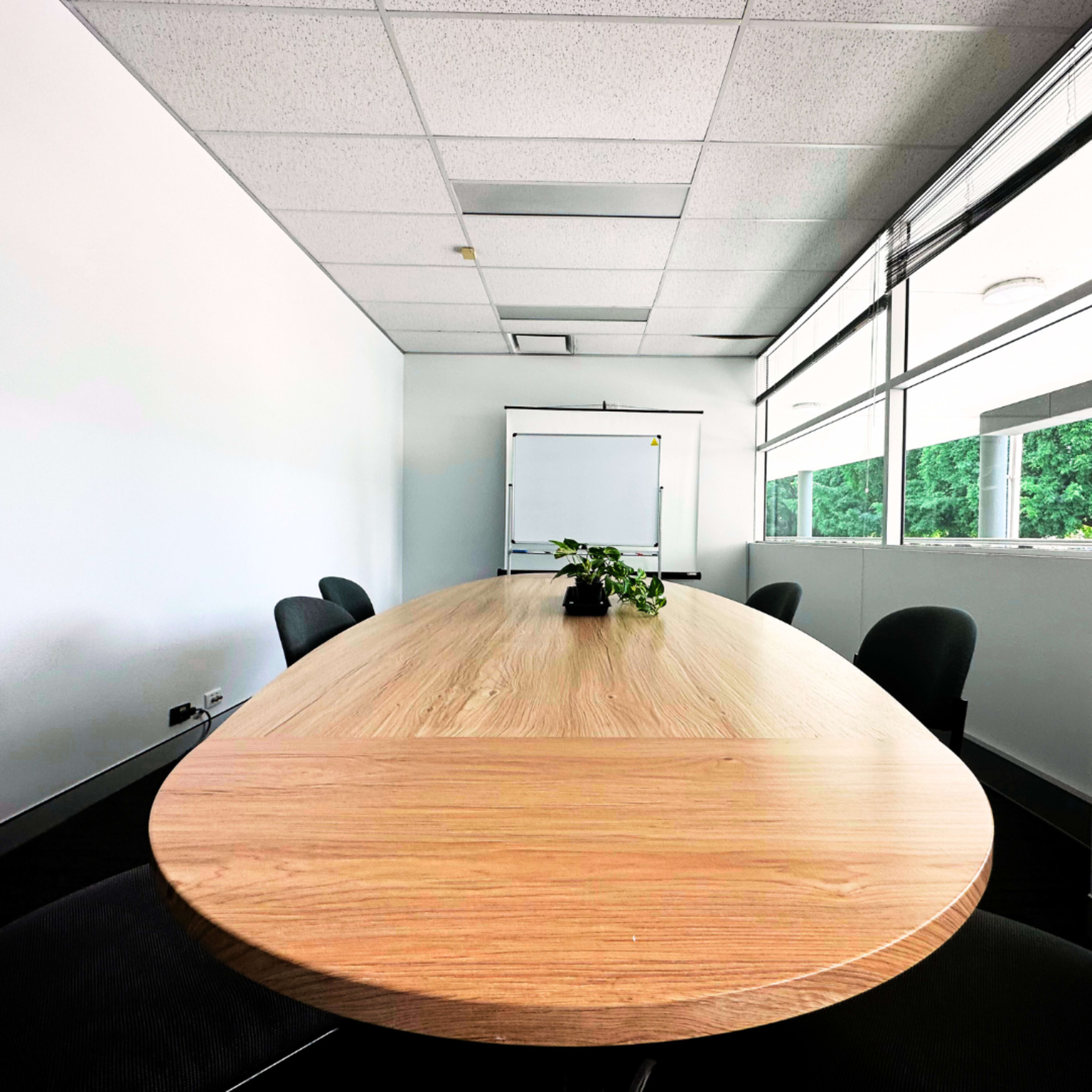 A large, oval wooden conference table is set in a bright meeting room, accompanied by black chairs and a whiteboard at the far end.
