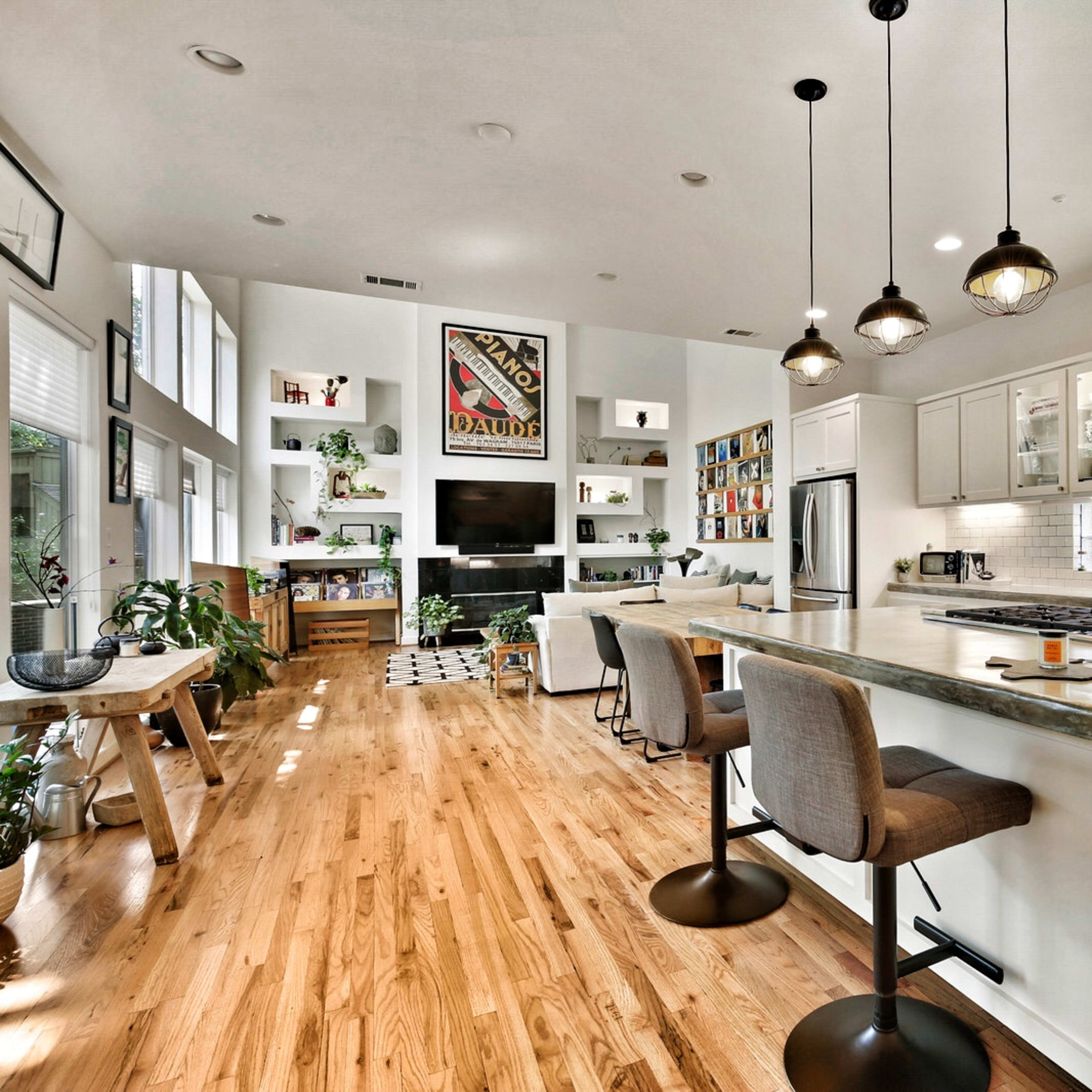 A modern kitchen features a central island with stools, white cabinets, wooden flooring, and a living area in the background with shelves and a television.
