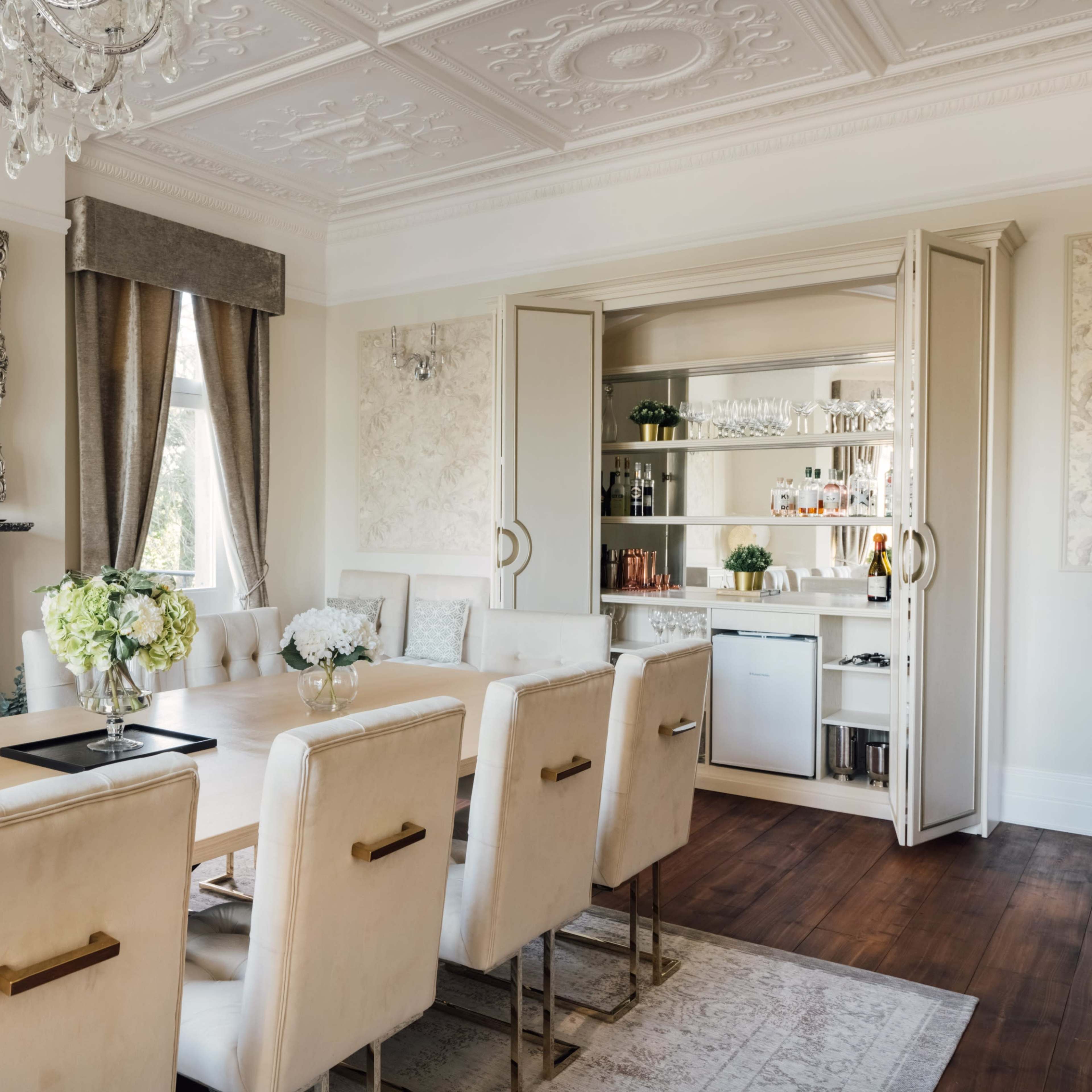 A formal dining room featuring a long wooden table surrounded by upholstered chairs, with a chandelier and a mirrored cabinet displaying glassware in the background.