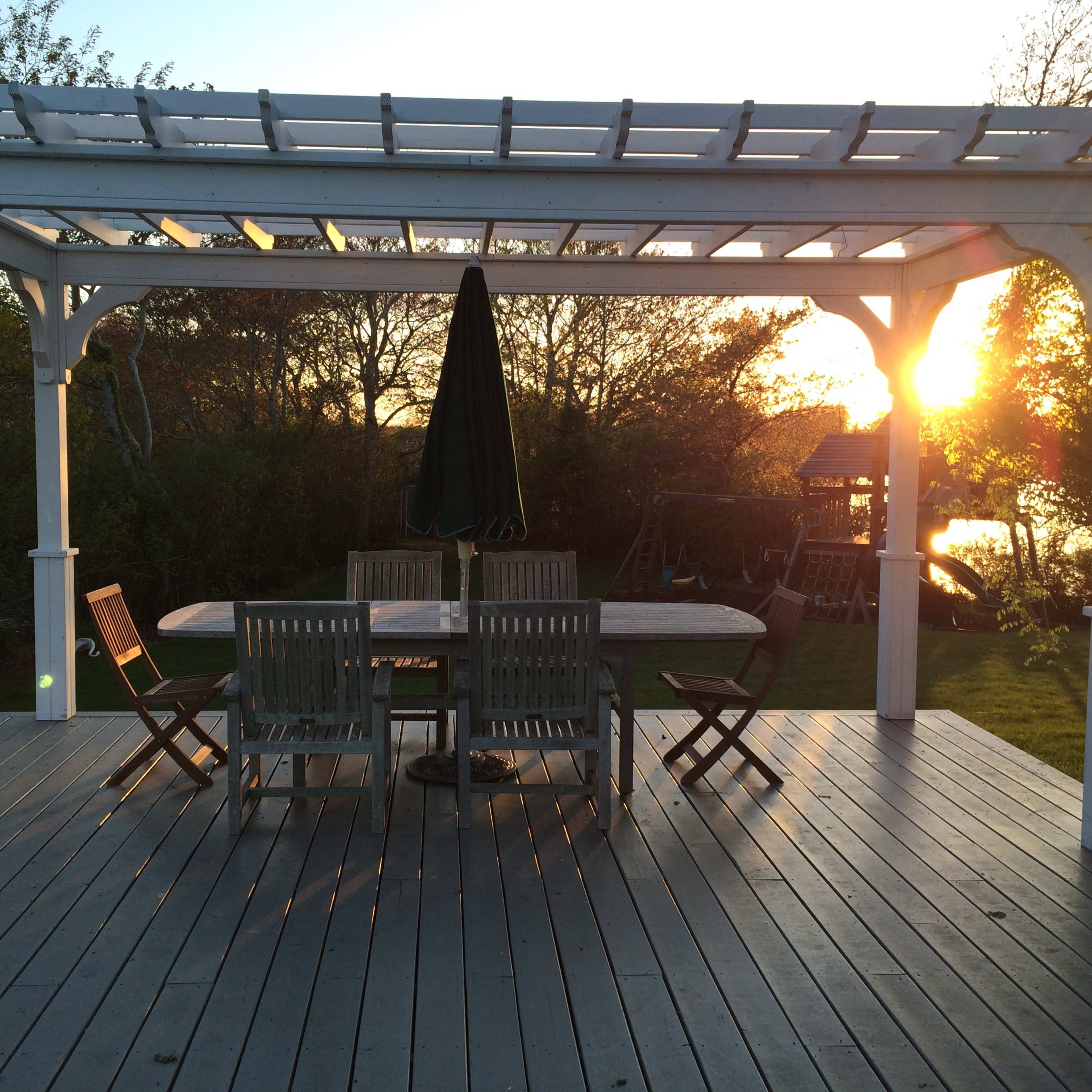 A white pergola with a shaded table and chairs sits on a wooden deck, framed by a sunset over a tranquil landscape.
