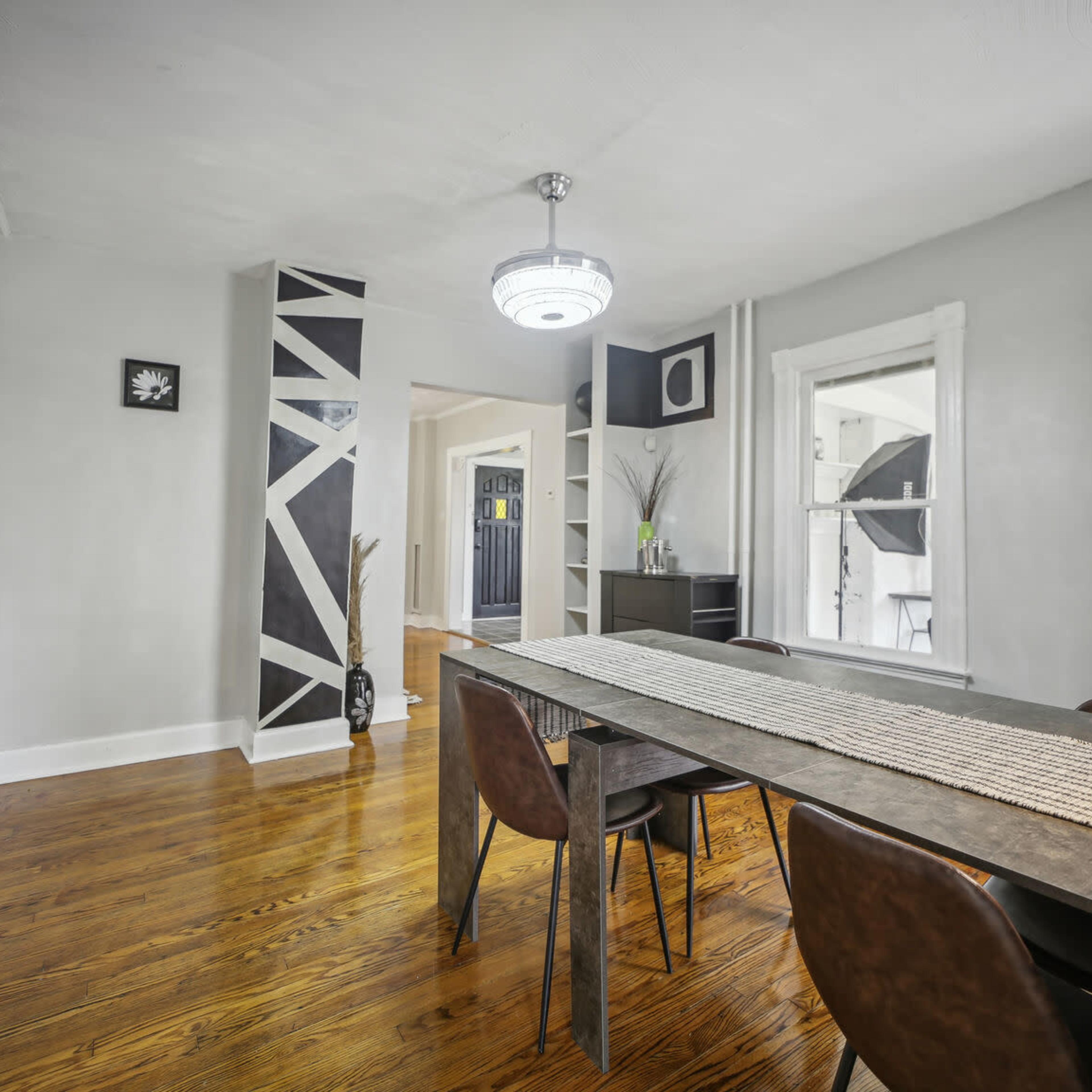 A dining area features a long table with chairs, a geometric accent wall, and a modern light fixture.
