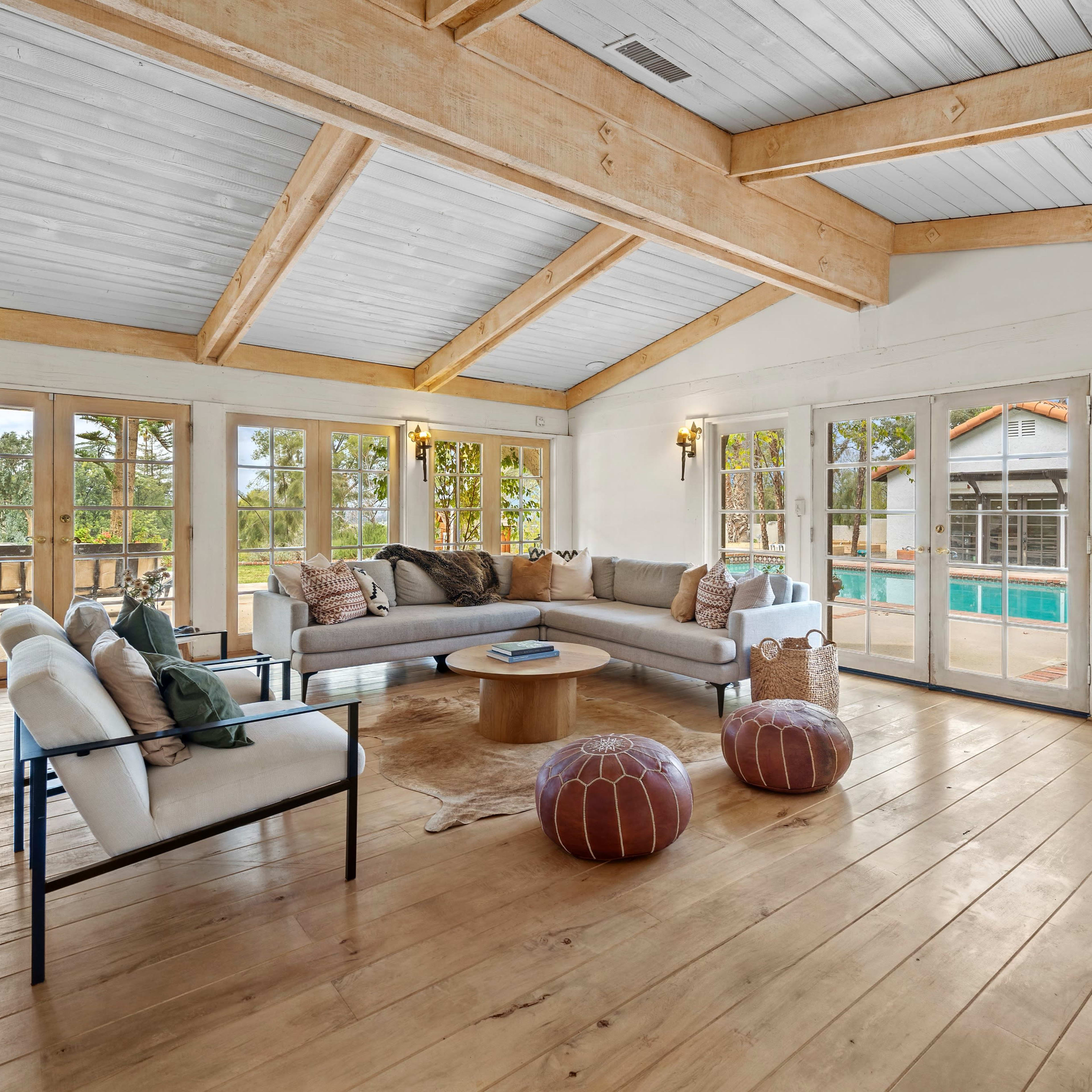 A spacious living room features large windows, wooden beams, and a view of a pool area outside.