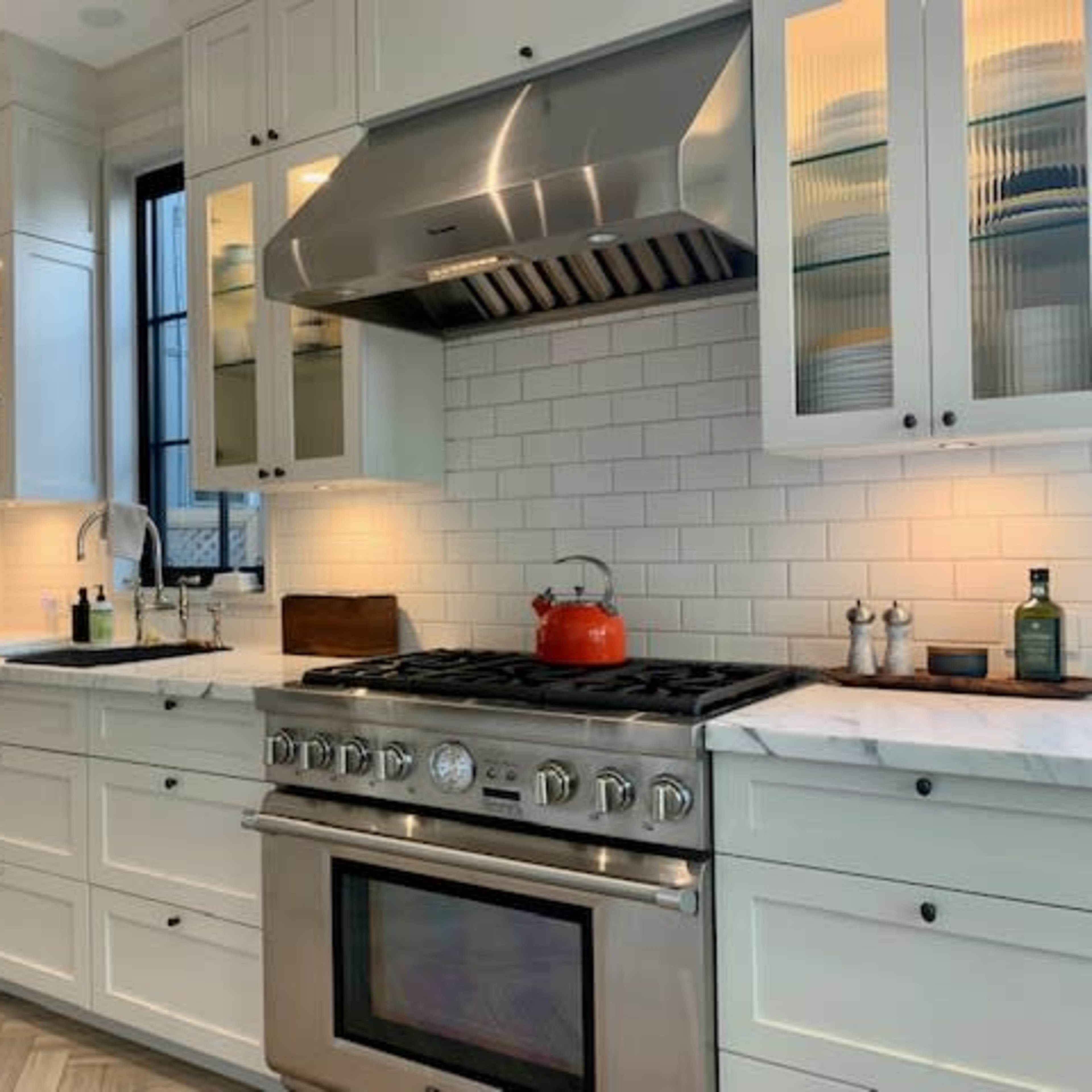 A modern kitchen with white cabinets, a large gas stove with a red kettle, and glass-fronted upper cabinets displaying dishware.