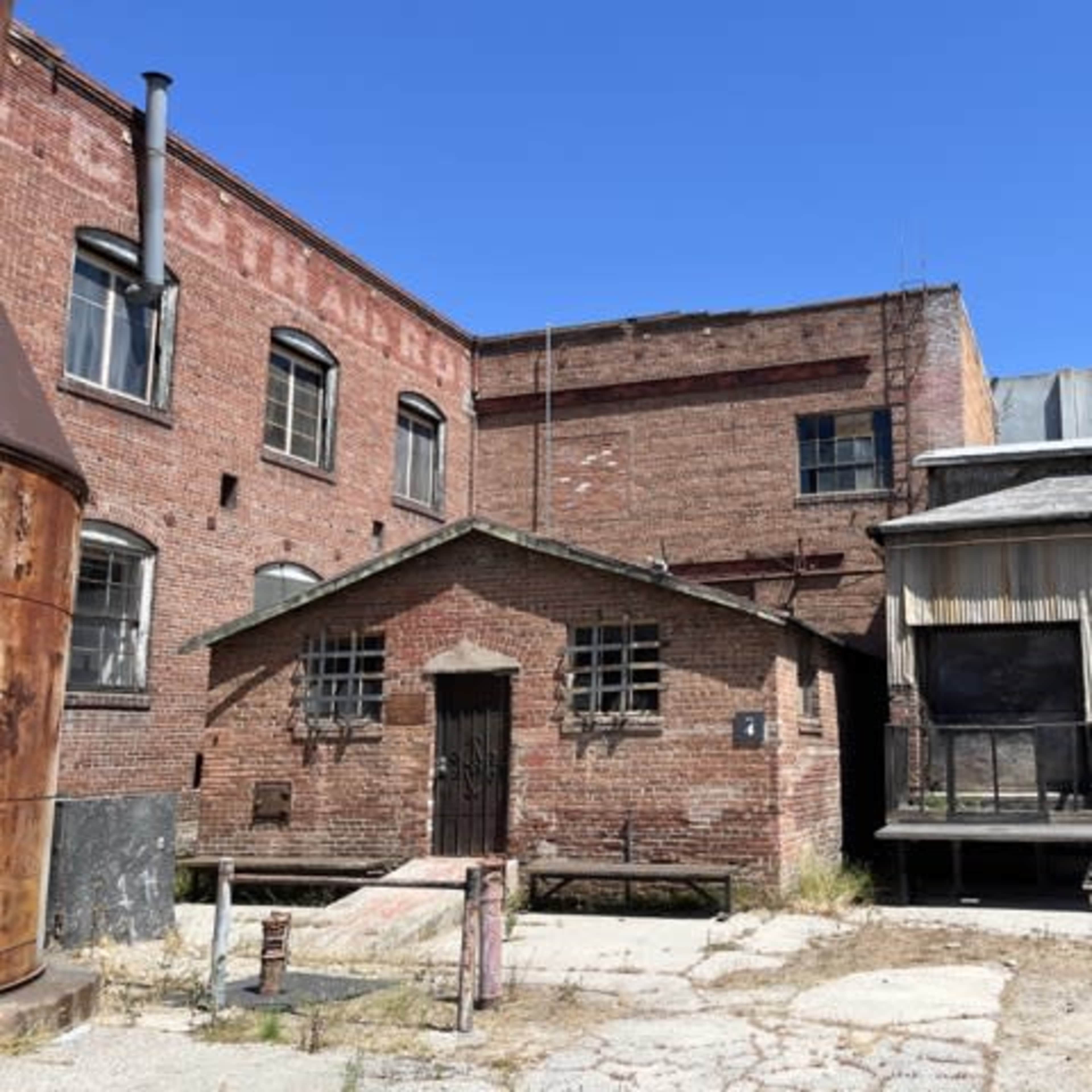 A collection of dilapidated brick and metal buildings in an overgrown industrial area under a clear blue sky.