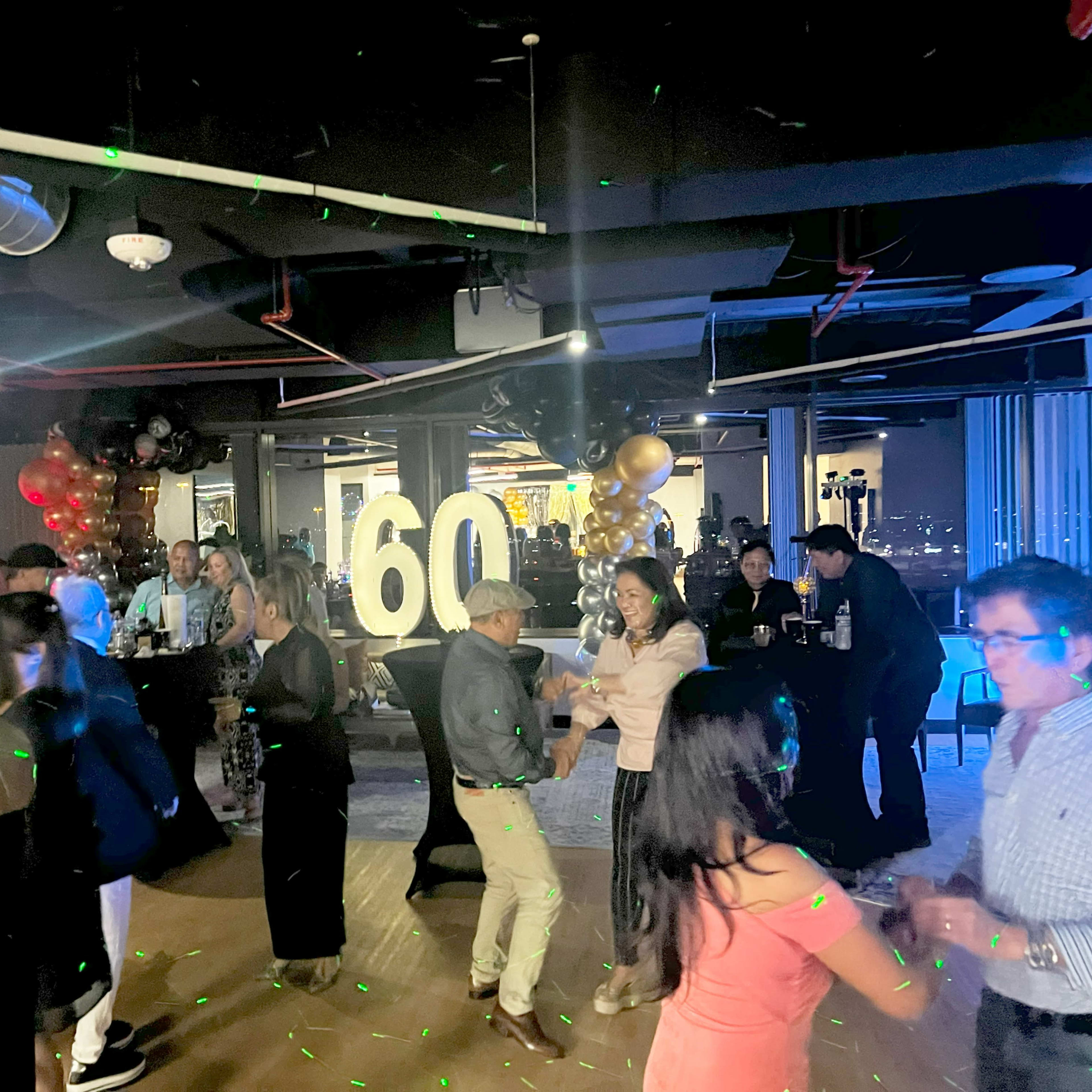 A lively party is taking place in an office space, featuring a large "60" centerpiece and guests dancing amid colorful lights.