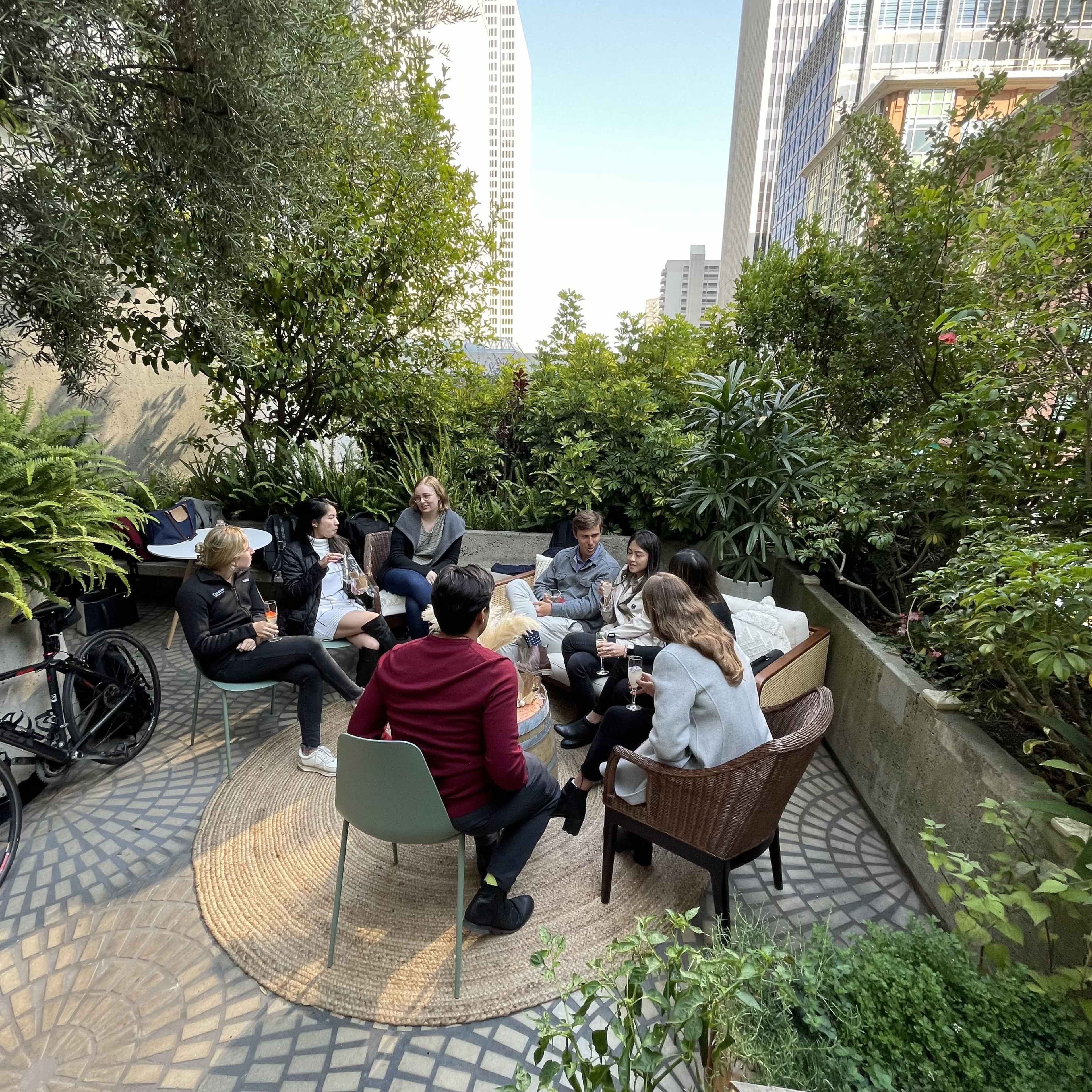 A group of six people sits in a landscaped outdoor space surrounded by greenery, engaging in conversation.