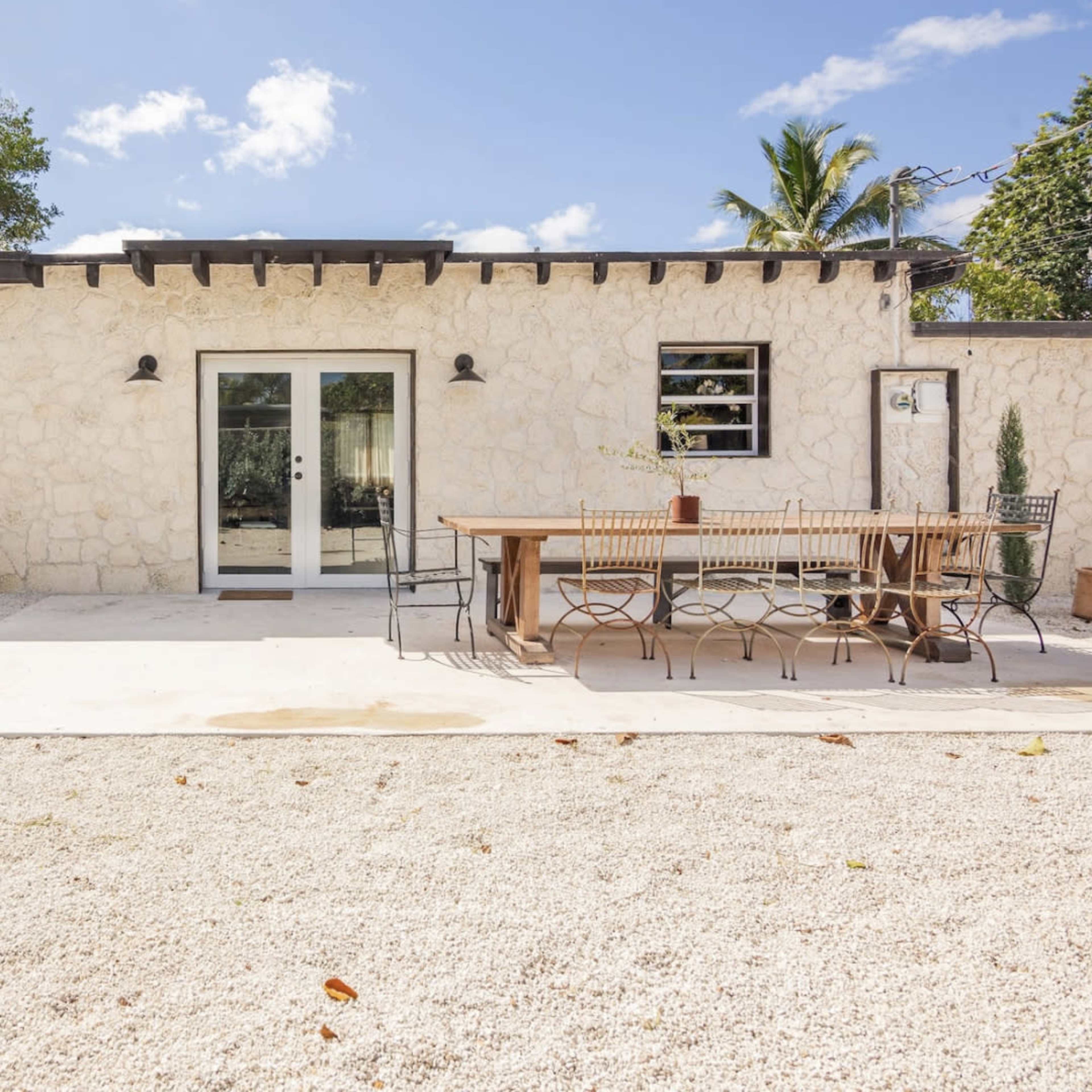 A stone-clad building features large glass doors opening to a patio with a long wooden dining table and chairs, surrounded by gravel and greenery.