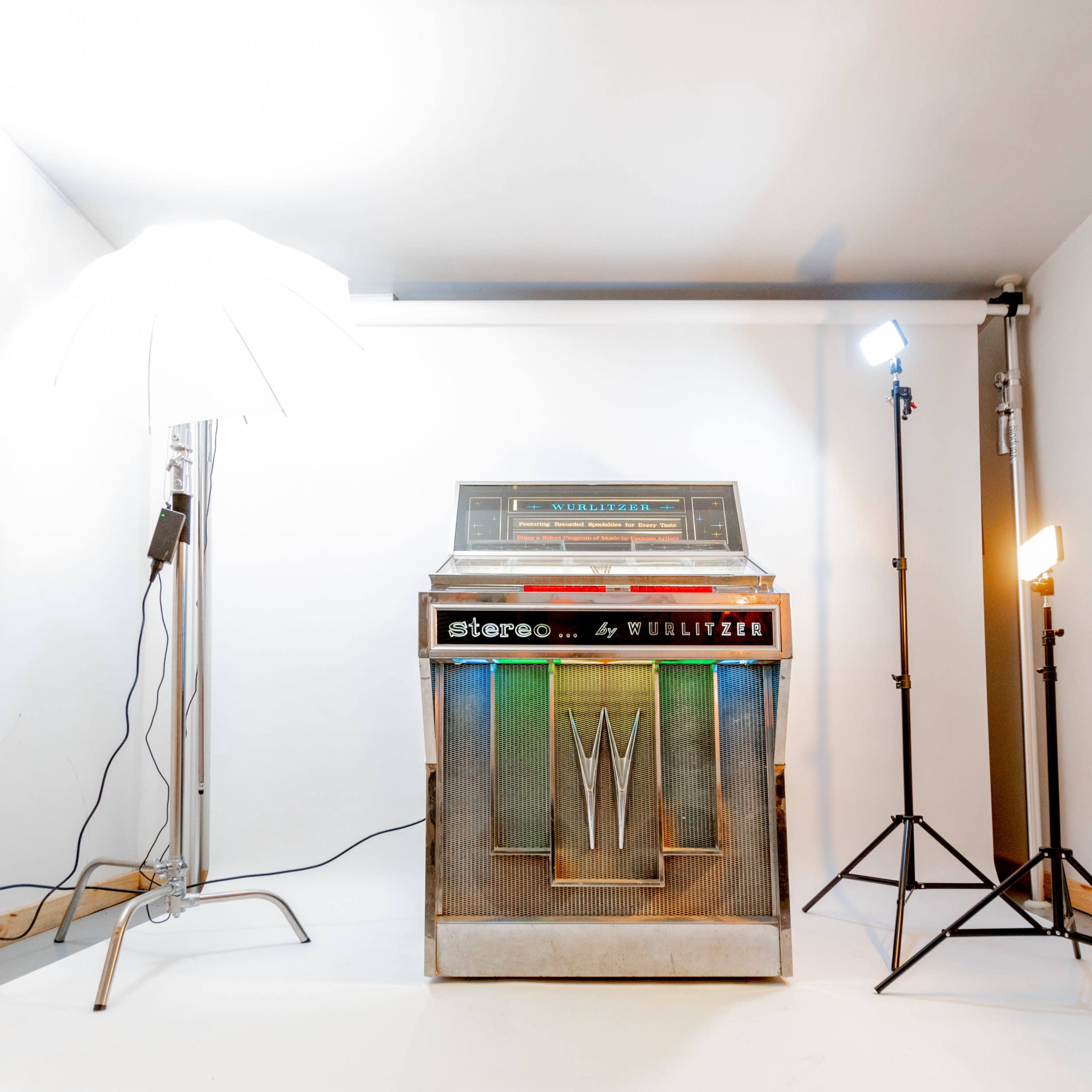 A vintage jukebox sits in a well-lit studio space, flanked by two light stands and an umbrella softbox.