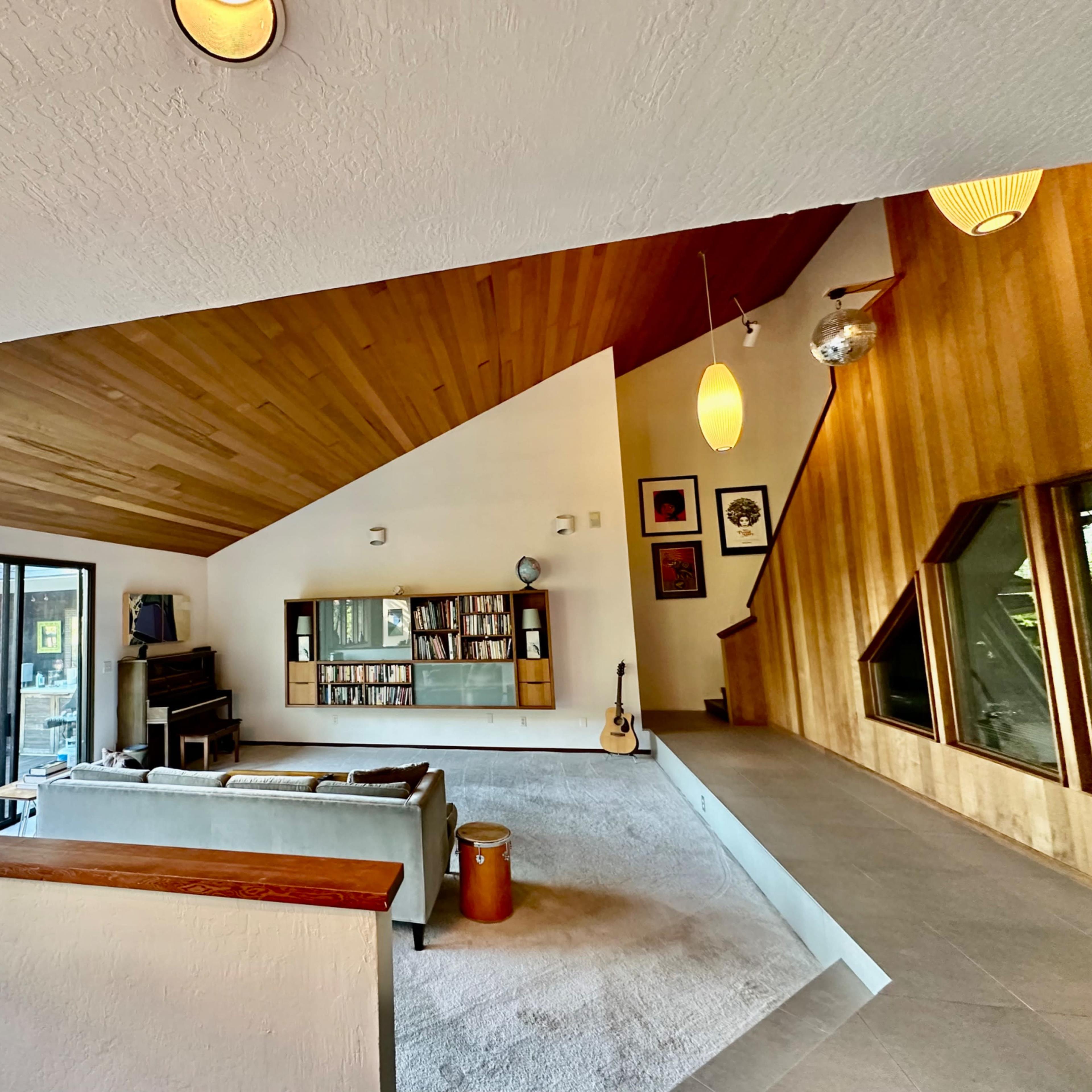 A modern living room features a sloped wooden ceiling, large windows, a bookshelf, and a seating area with a view.