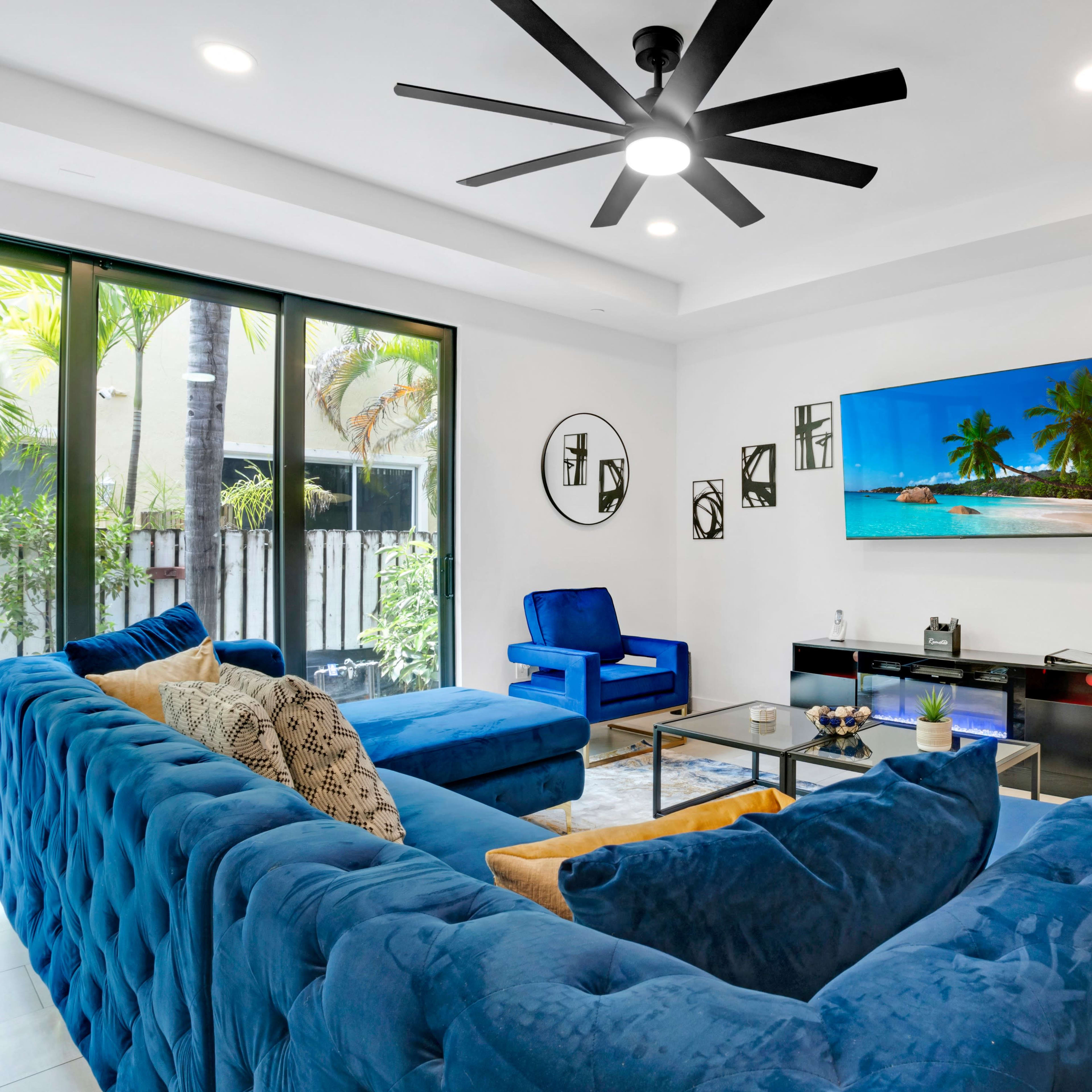 A spacious living room features a large blue sectional sofa, a wall-mounted television, and modern decor with palm trees visible through large windows.