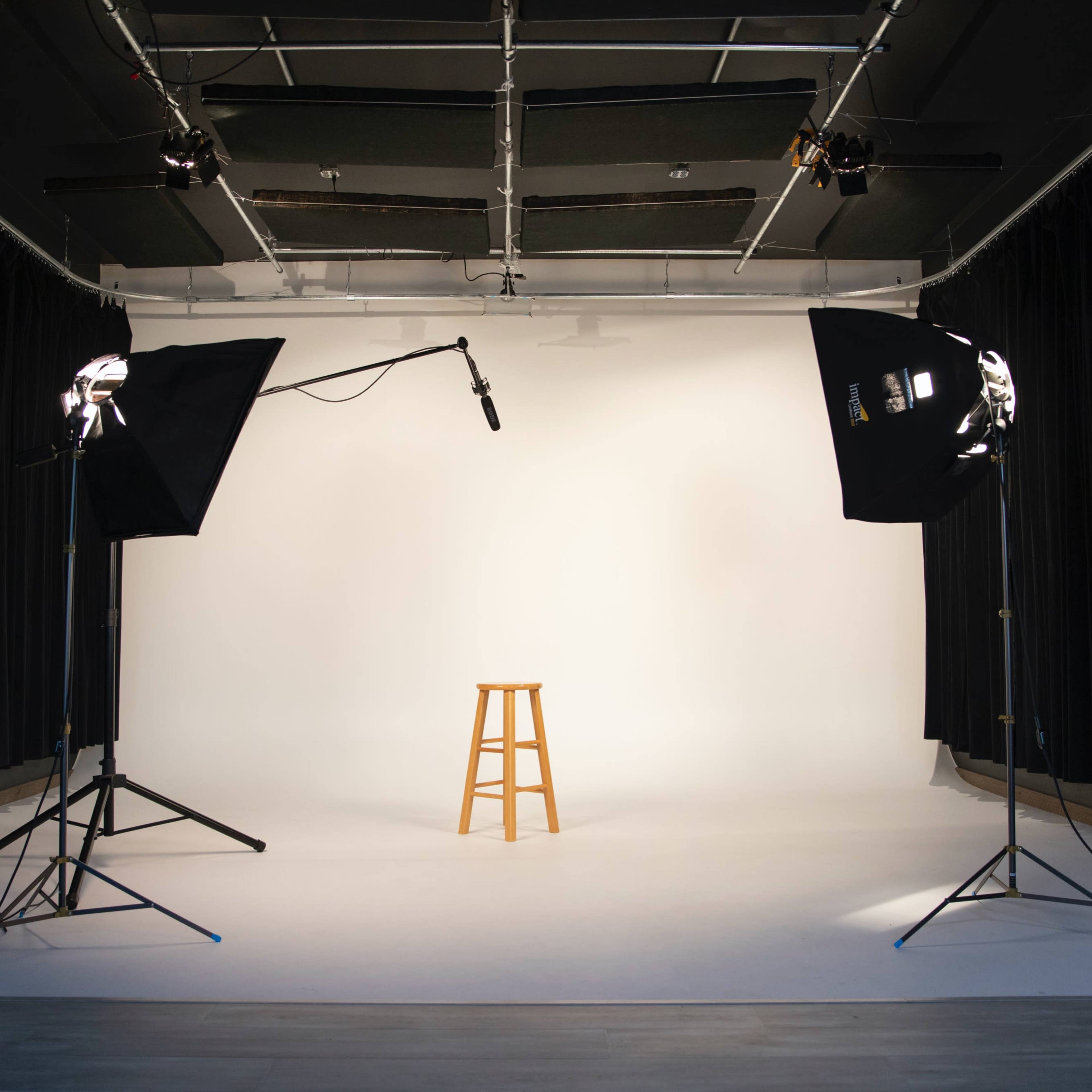 A wooden stool is positioned in the center of a well-lit photography studio with two softbox lights and a microphone overhead.