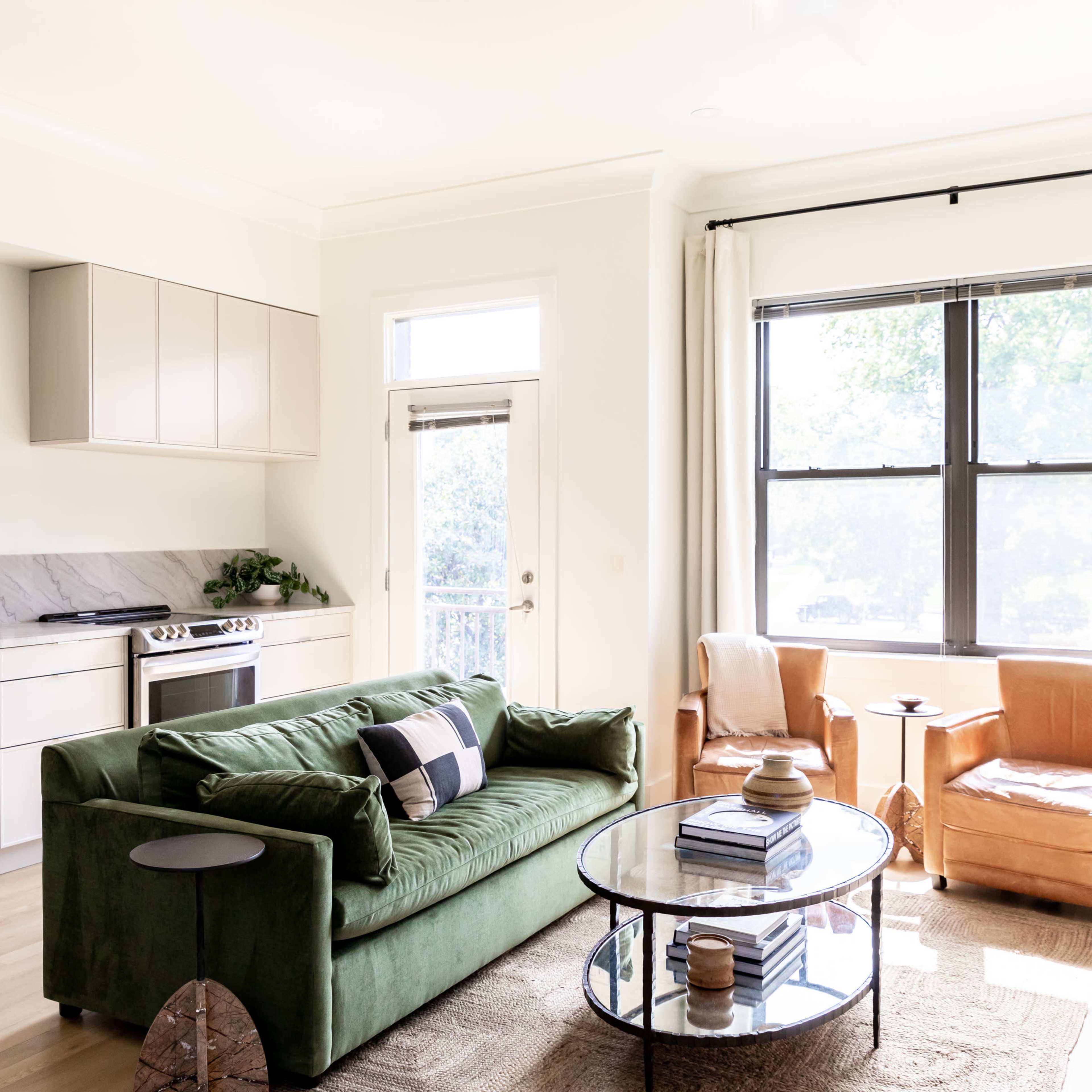 A modern living room features a green sofa, a round glass coffee table, and two orange armchairs, with a kitchen area visible in the background.