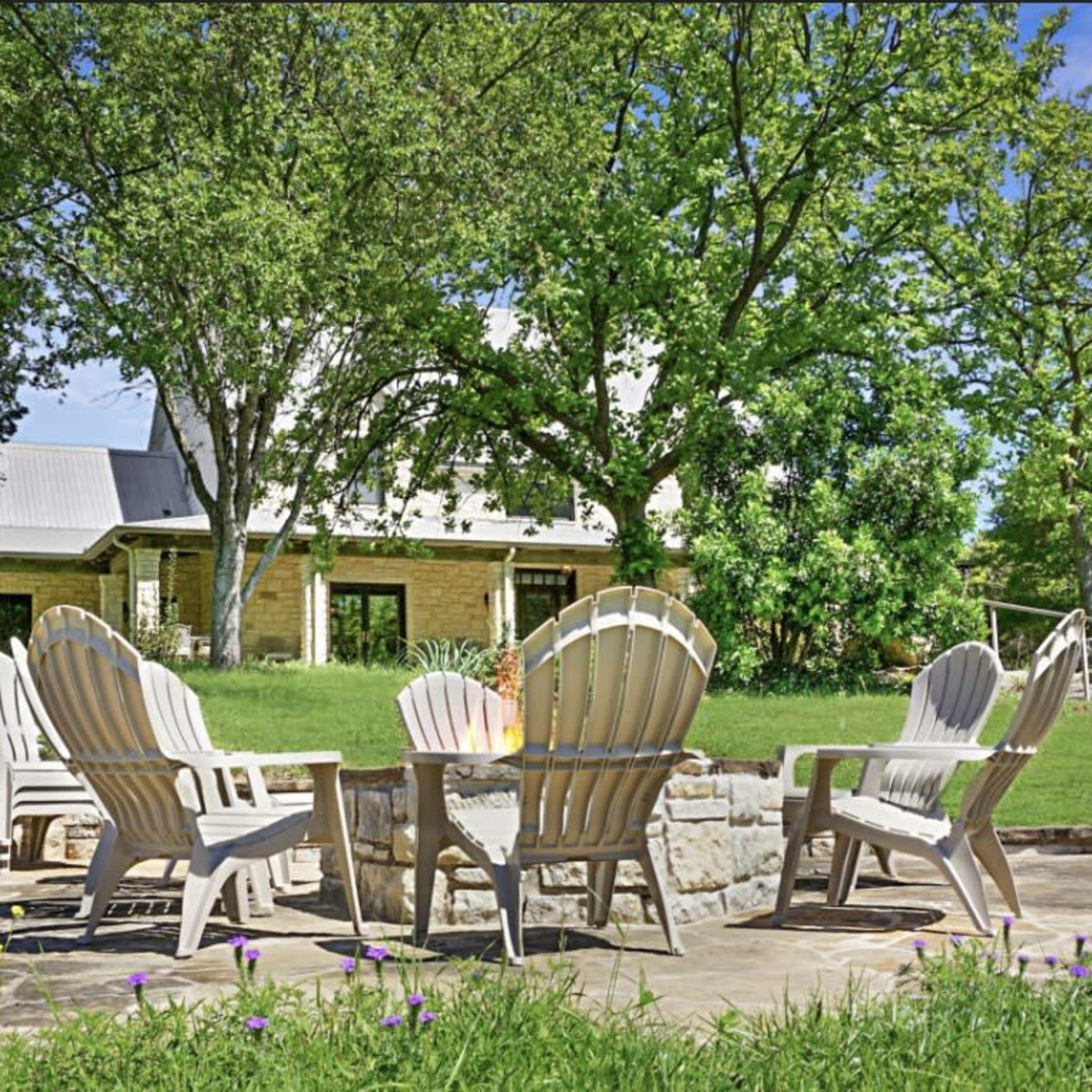 A stone patio features five white Adirondack chairs arranged around a fire pit, with a large house and lush greenery in the background.