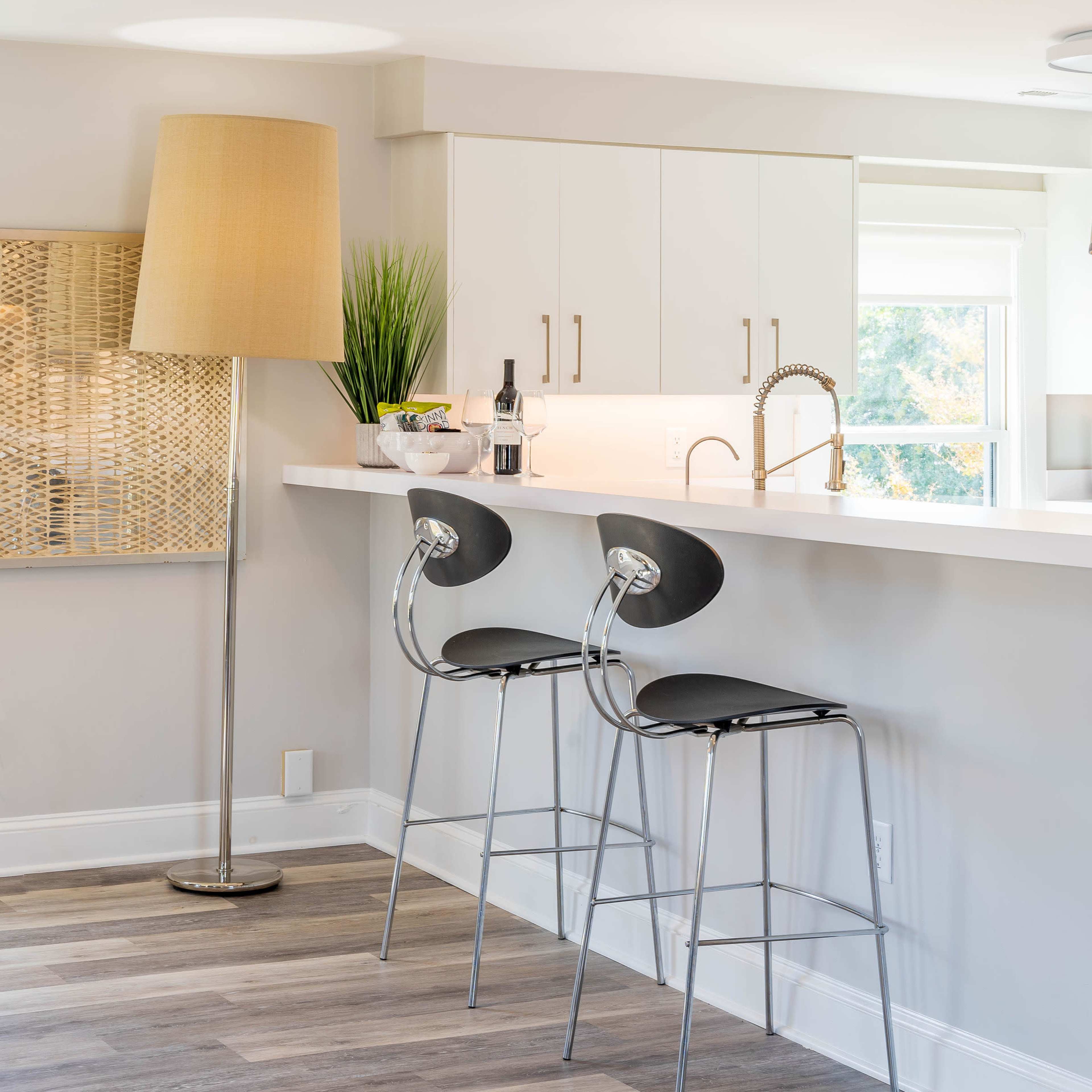 A modern kitchen features a white countertop with two black bar stools, a tall floor lamp, and a small plant, alongside cabinetry and a bottle of wine.