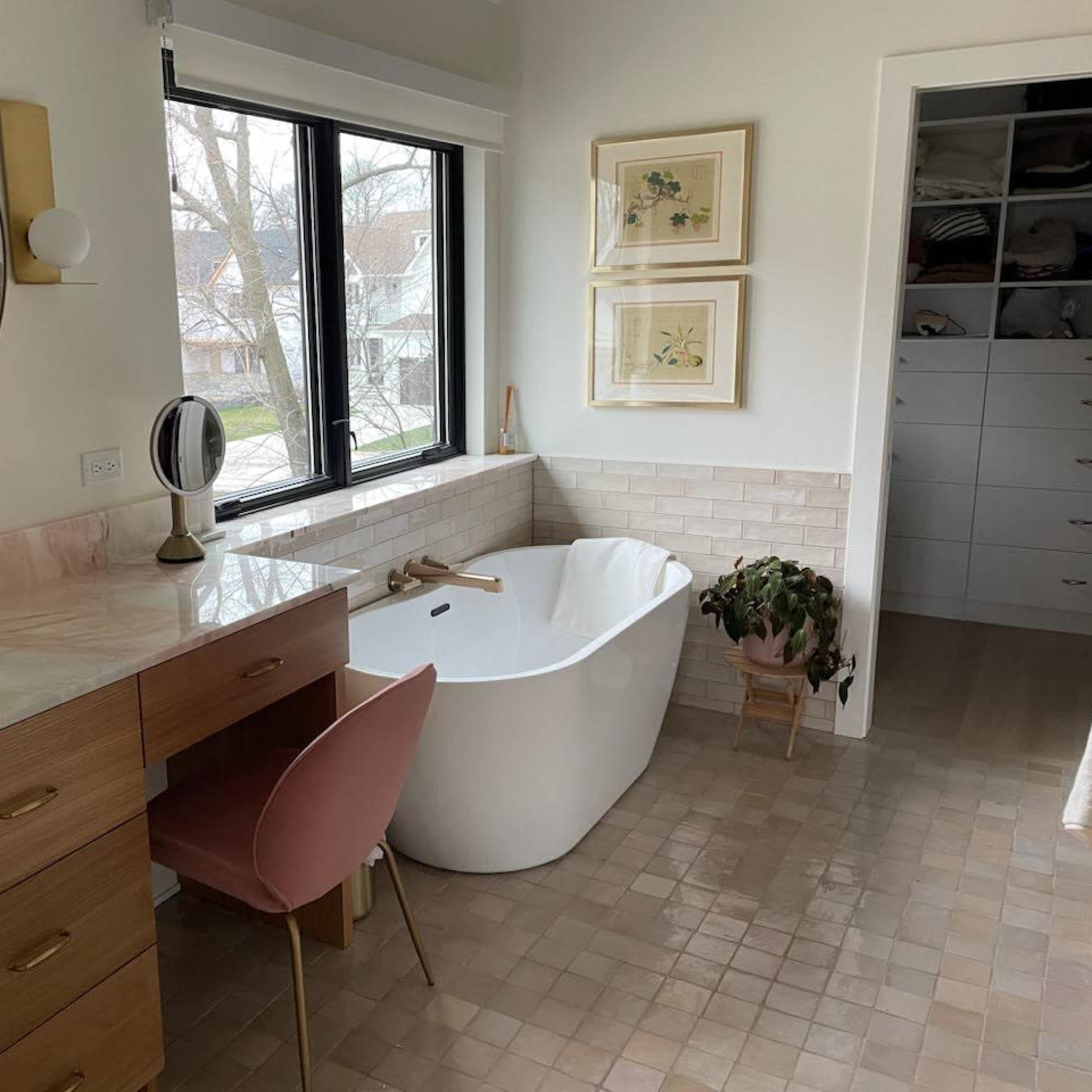A modern bathroom featuring a freestanding bathtub, a wooden vanity with a pink chair, and large windows that let in natural light.