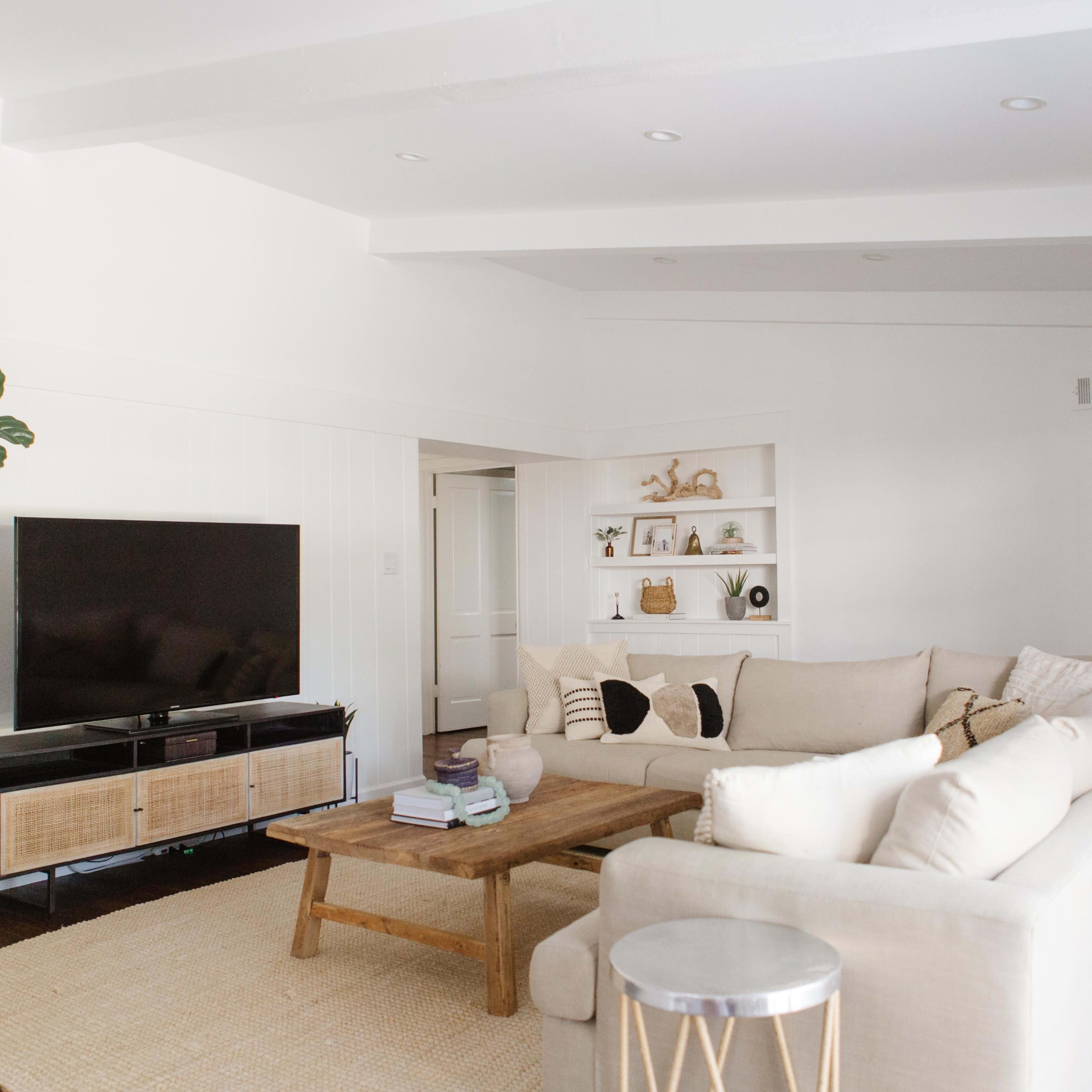 A spacious living room features a large sectional sofa, a coffee table, a television on a stand, and a potted plant, all arranged around a light-colored area rug.