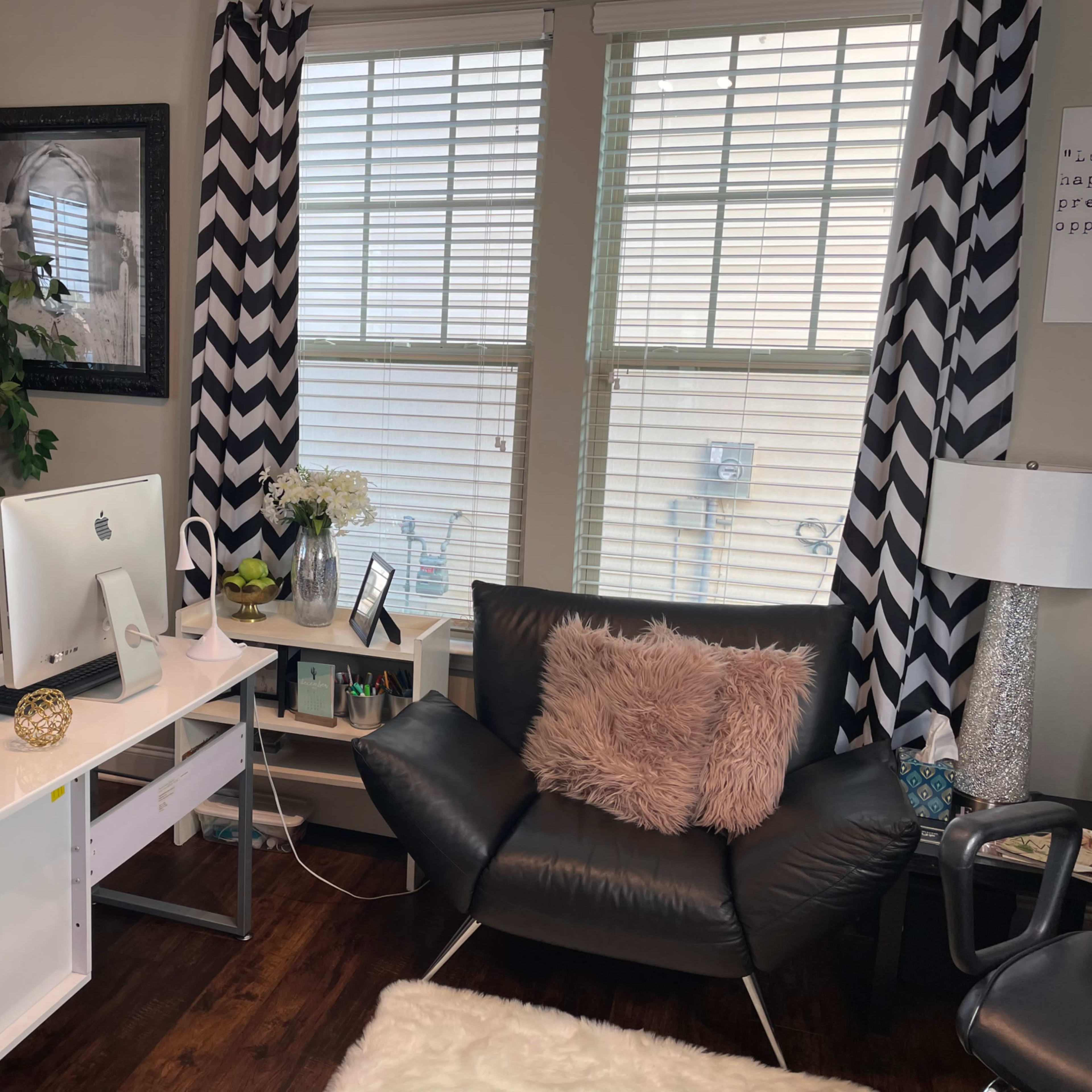 A modern office space features a white desk with an iMac, a black armchair with a pink pillow, and zigzag patterned curtains.