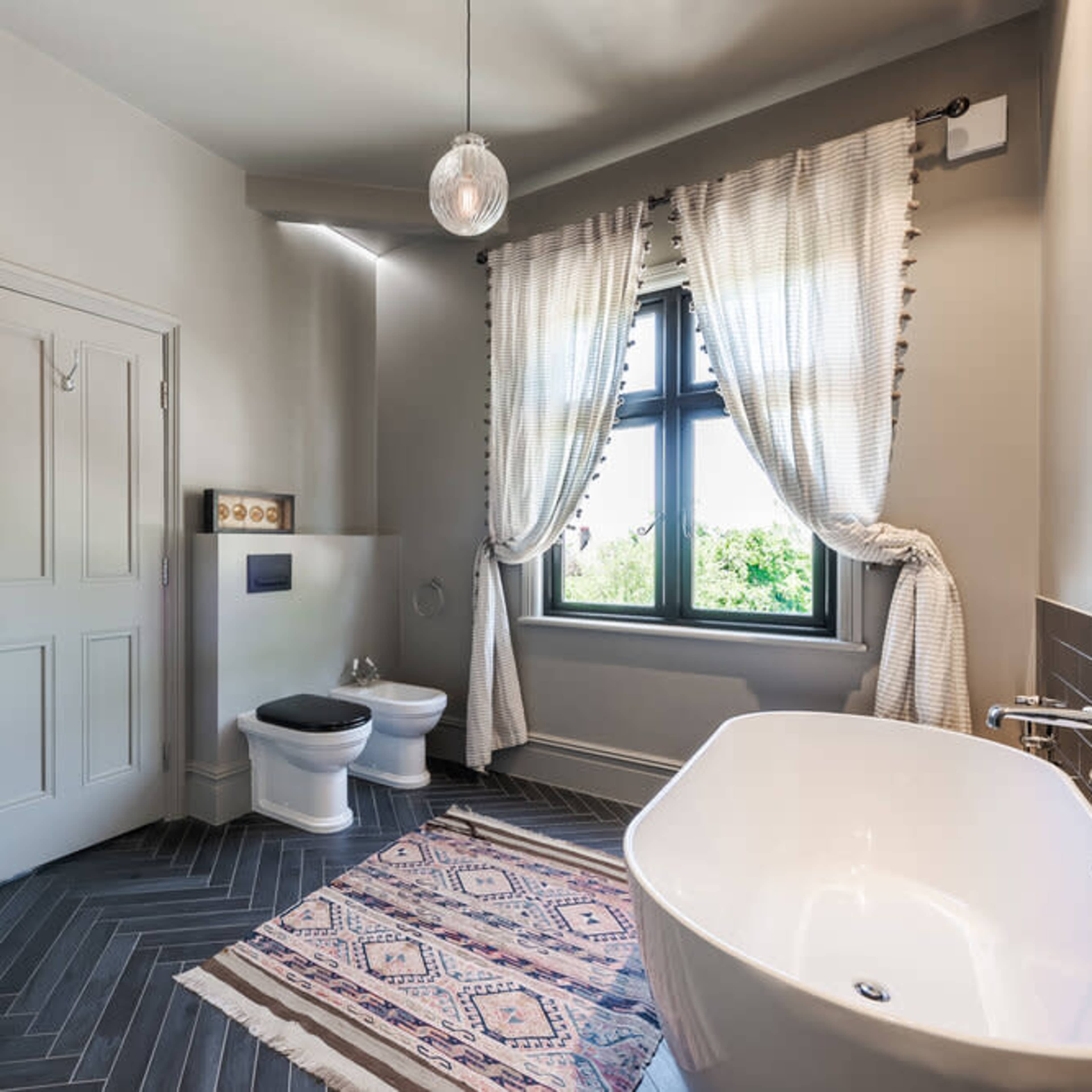 The bathroom features a freestanding bathtub, a wall-mounted toilet, and large windows with curtains, accompanied by a patterned rug on the herringbone floor.