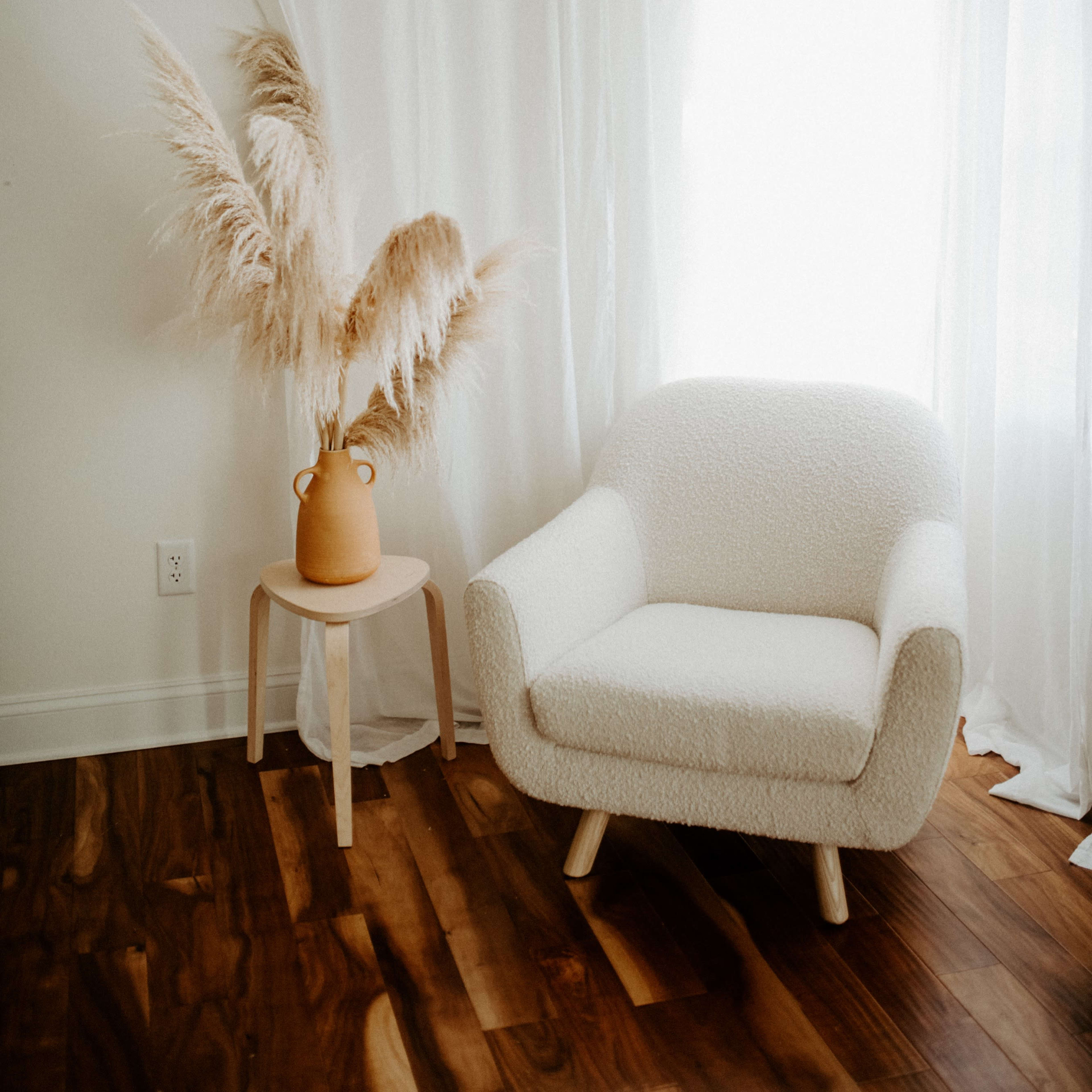A cozy chair sits beside a tall vase filled with pampas grass, accented by sheer white curtains and hardwood flooring.