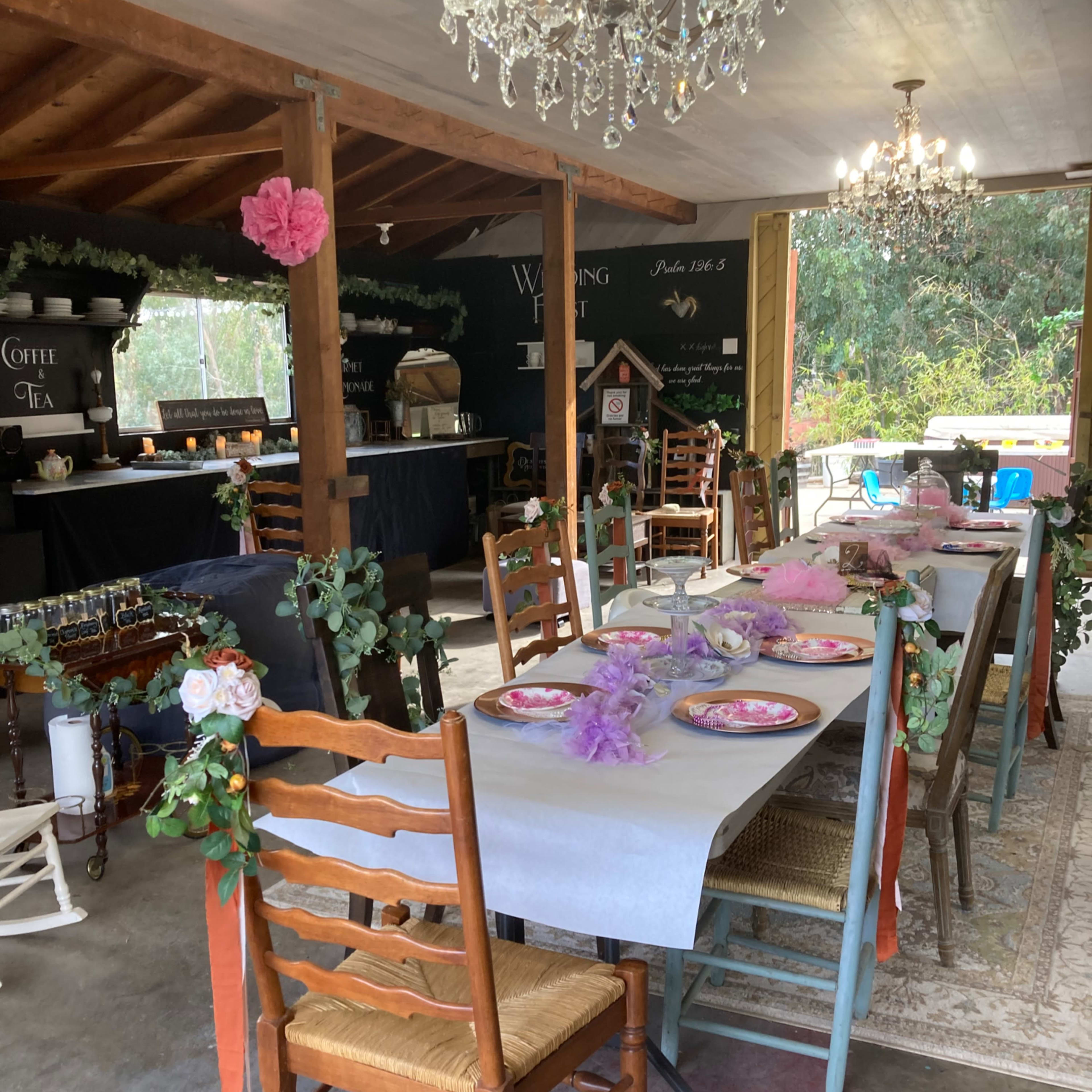 A decorated indoor space features tables set with plates and floral arrangements, surrounded by wooden chairs and chandeliers.