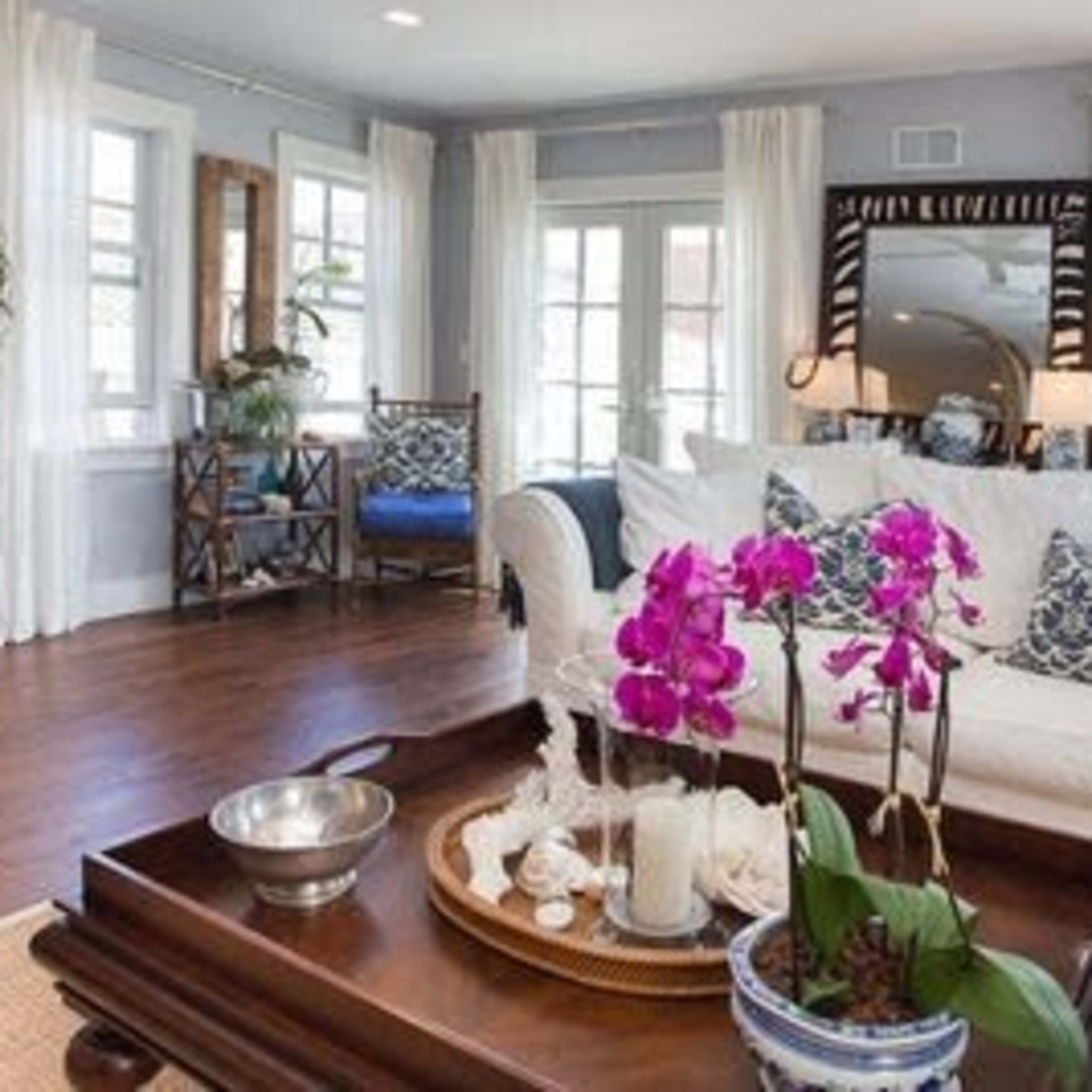 A cozy living room features a white sofa with colorful throw pillows, a wooden coffee table adorned with an arrangement of orchids, and large windows dressed with light curtains.