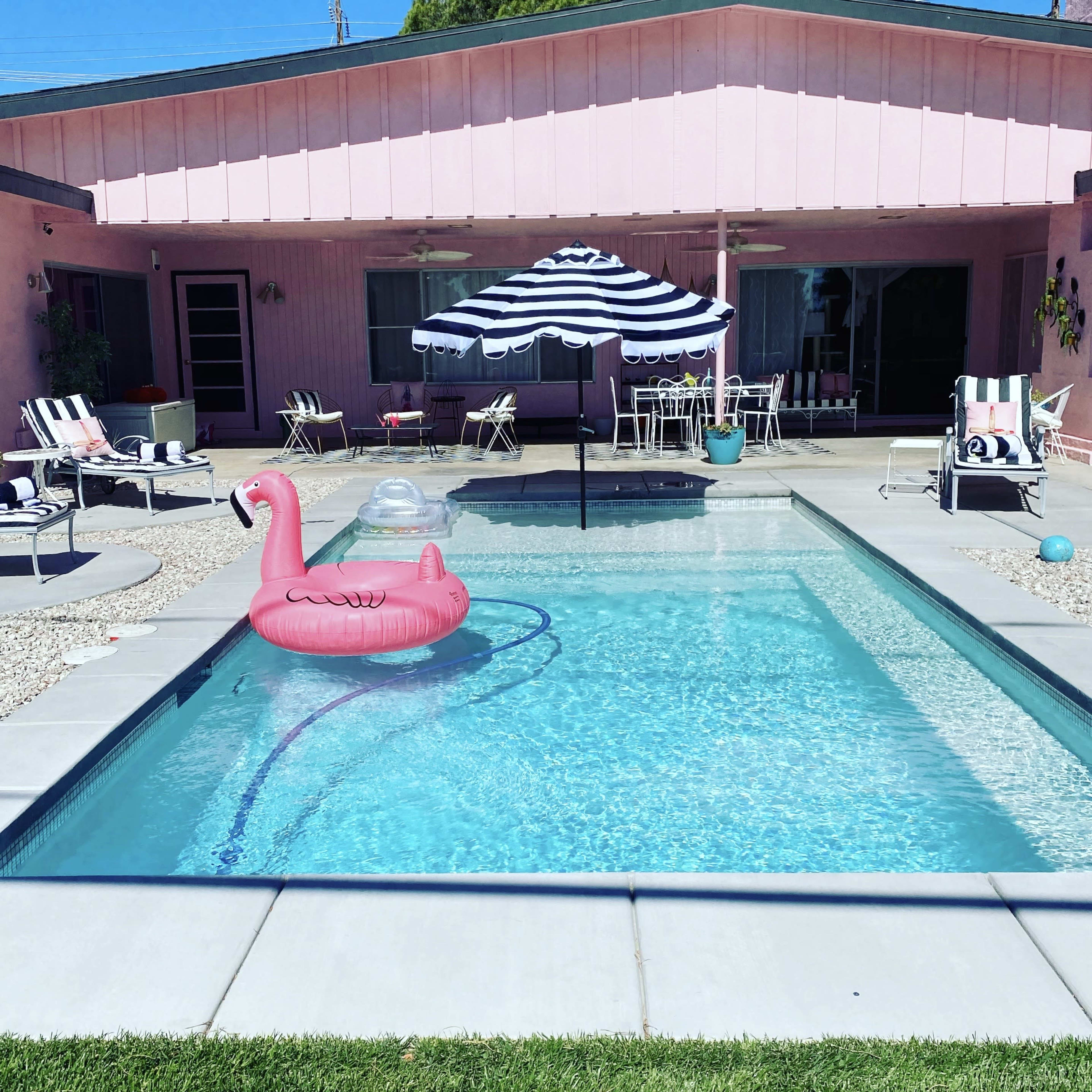 A pink flamingo float sits in a swimming pool surrounded by a patio with striped umbrellas and lounge chairs.