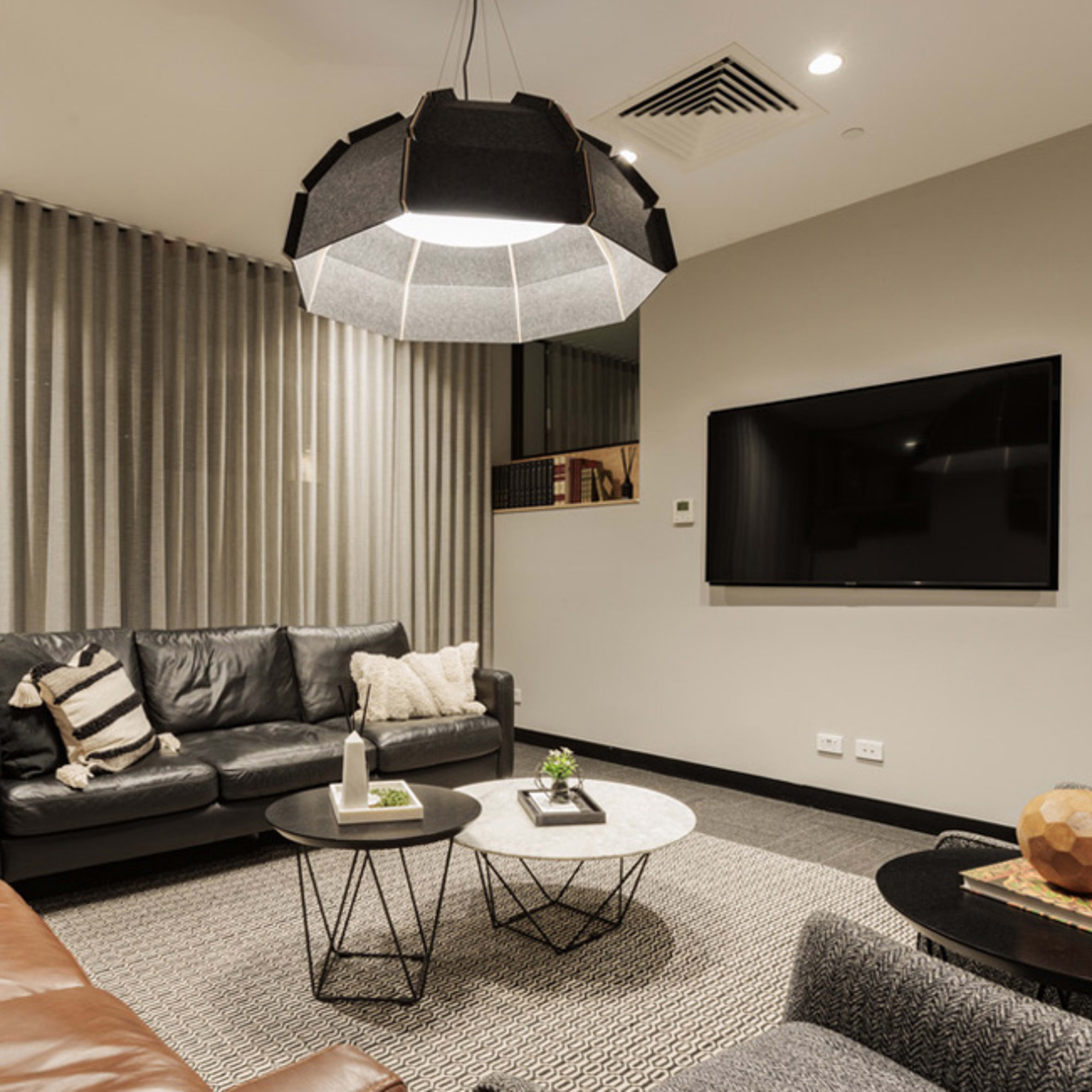 A modern living room featuring a black leather sofa, a brown leather couch, a large pendant light, and two coffee tables placed on a textured rug, with a television mounted on the wall.