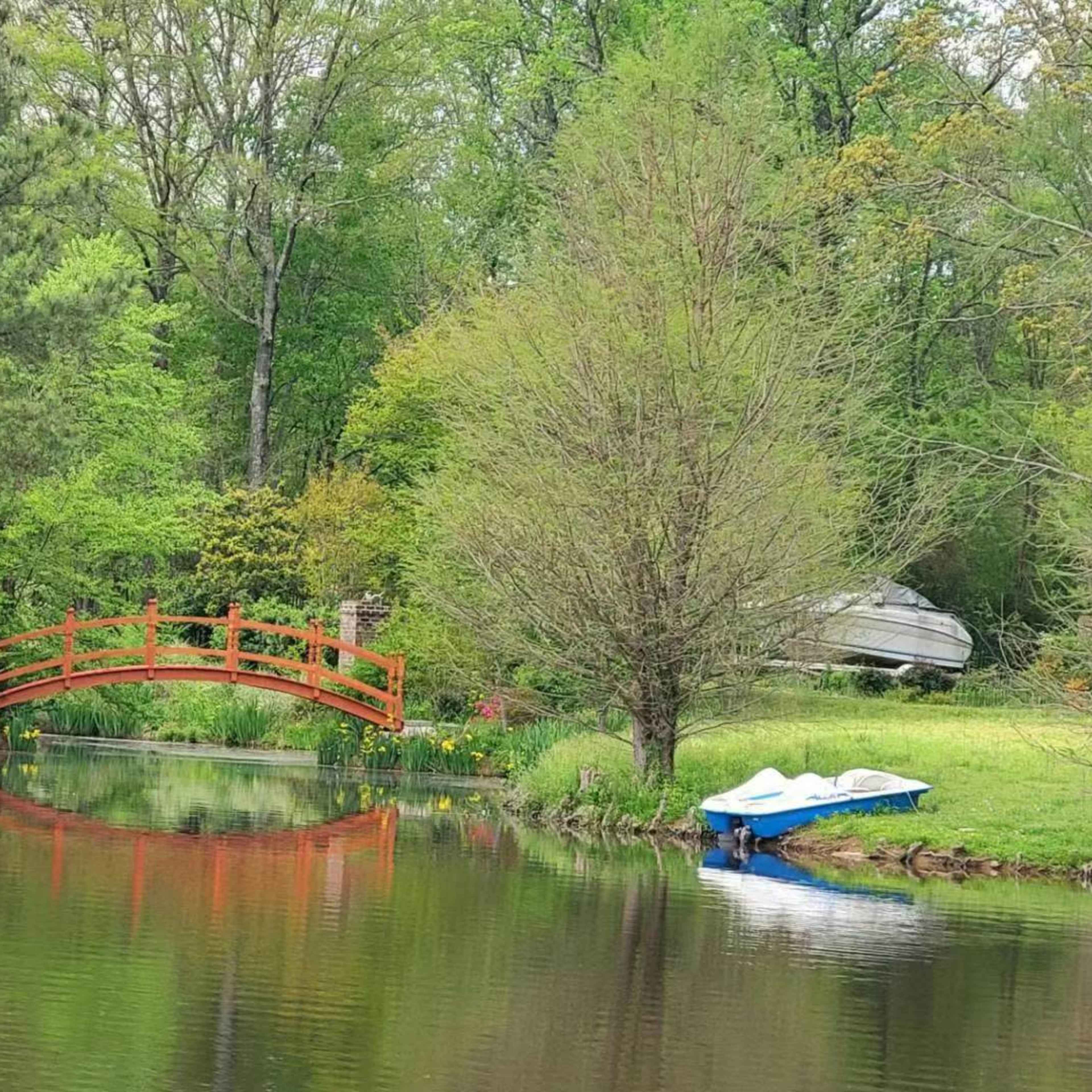 A red wooden bridge spans a calm pond surrounded by greenery and colorful flowers, with a small blue boat moored nearby.