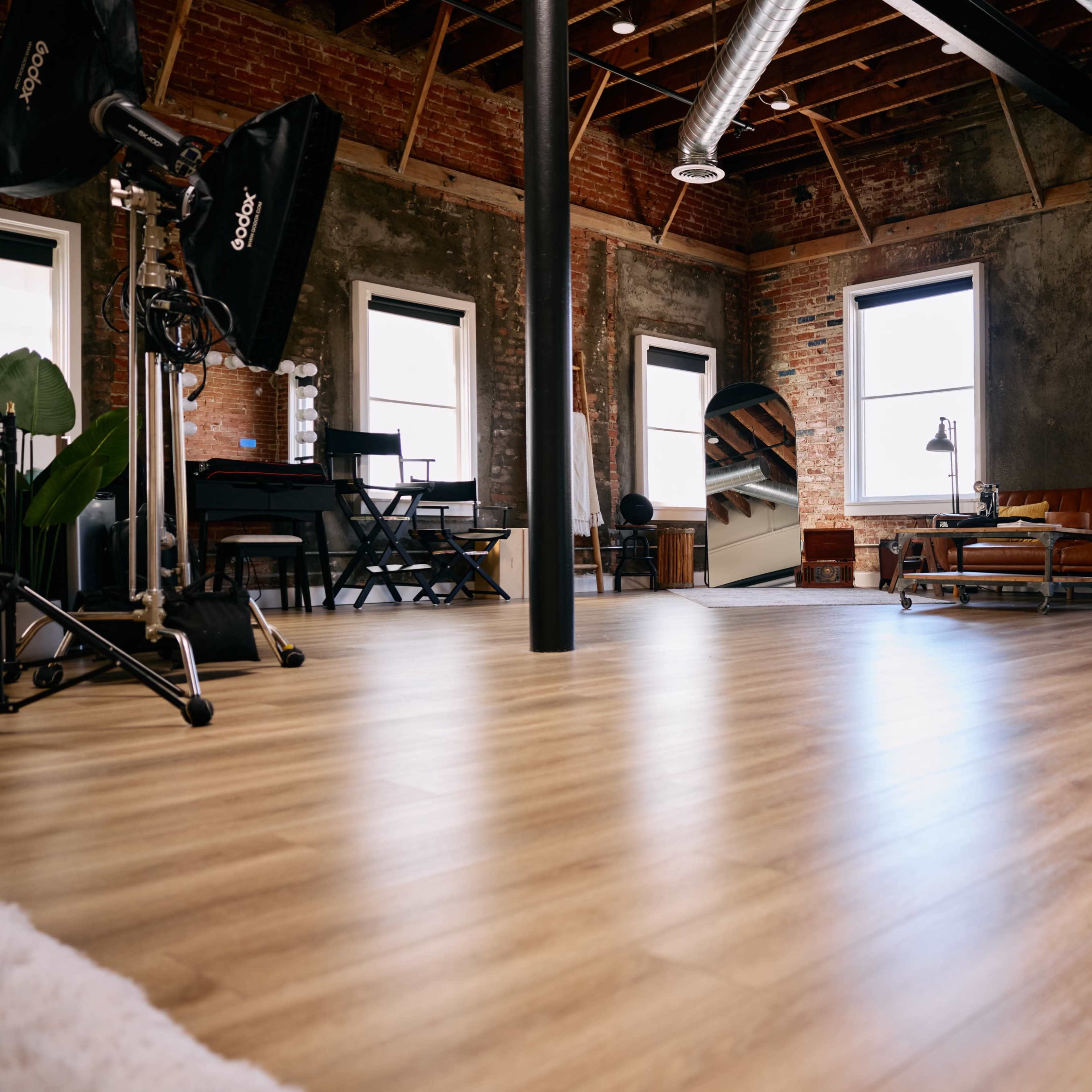 A spacious, retro-style studio with large windows, wooden flooring, and various furniture pieces, including a couch and a workspace.