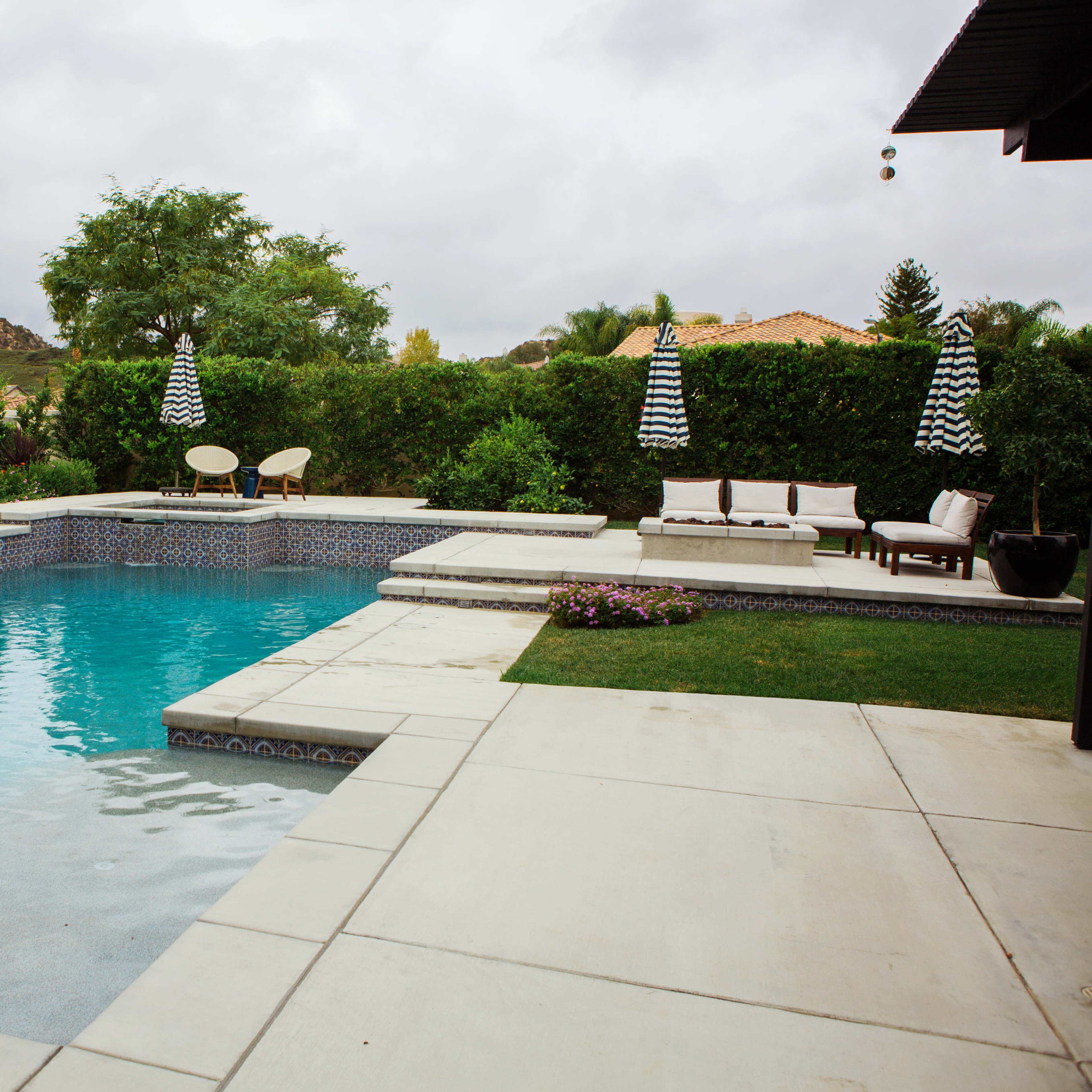 A swimming pool surrounded by concrete decking, with lounge chairs and striped umbrellas set in a landscaped backyard.
