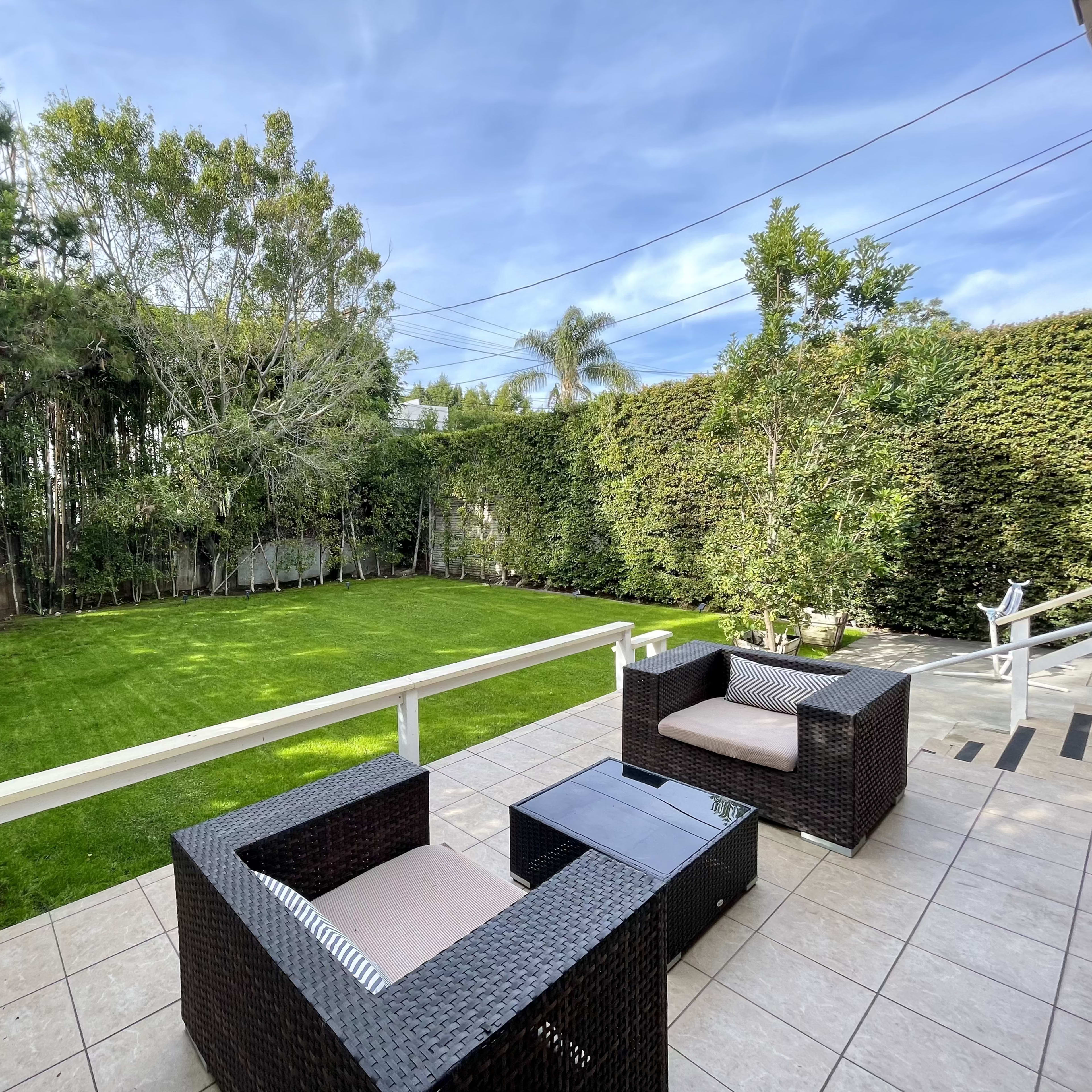 A neatly maintained backyard features a manicured lawn surrounded by tall hedges, with rattan chairs positioned on a tiled patio.