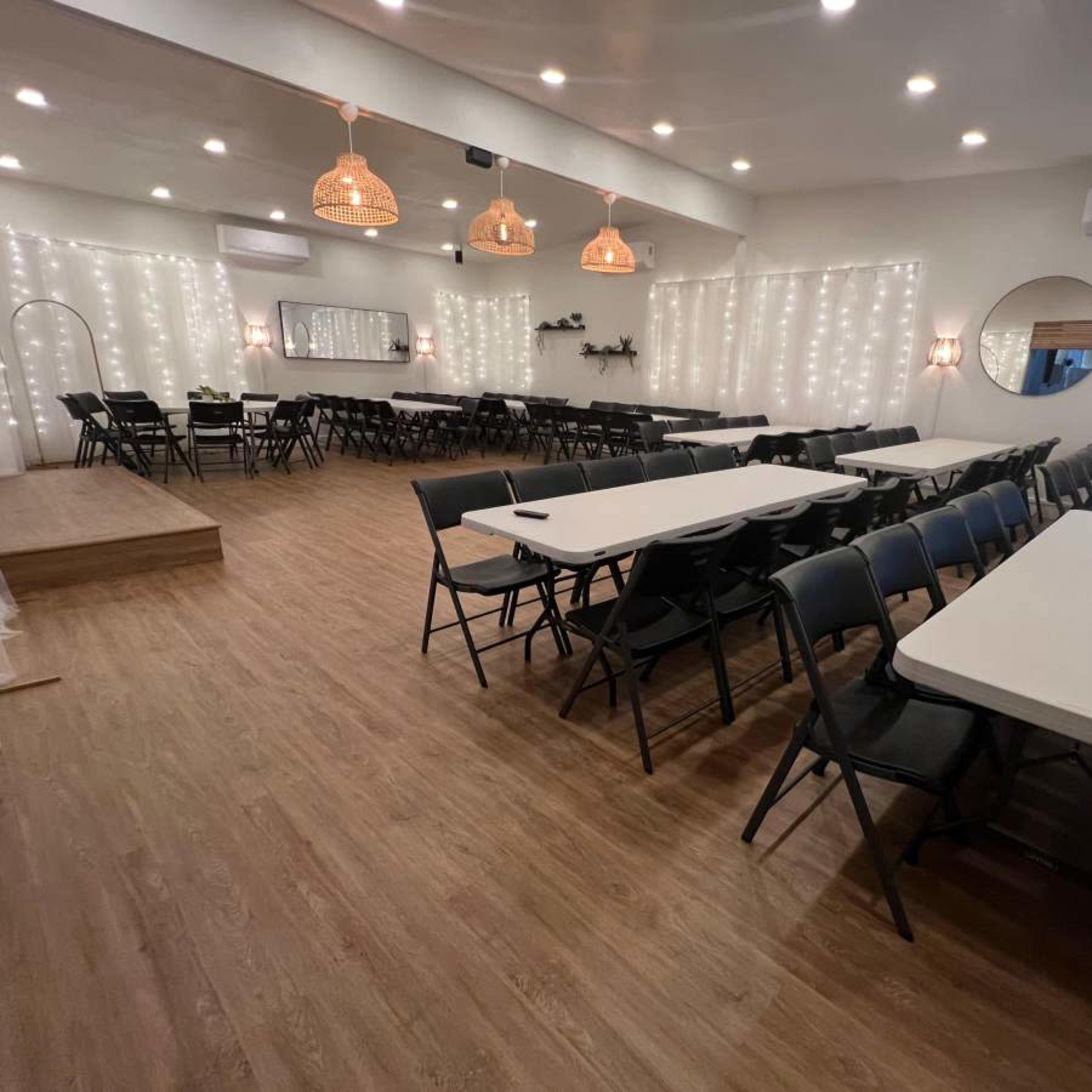 A spacious indoor venue arranged with multiple rectangular tables and black chairs, illuminated by string lights along the walls.