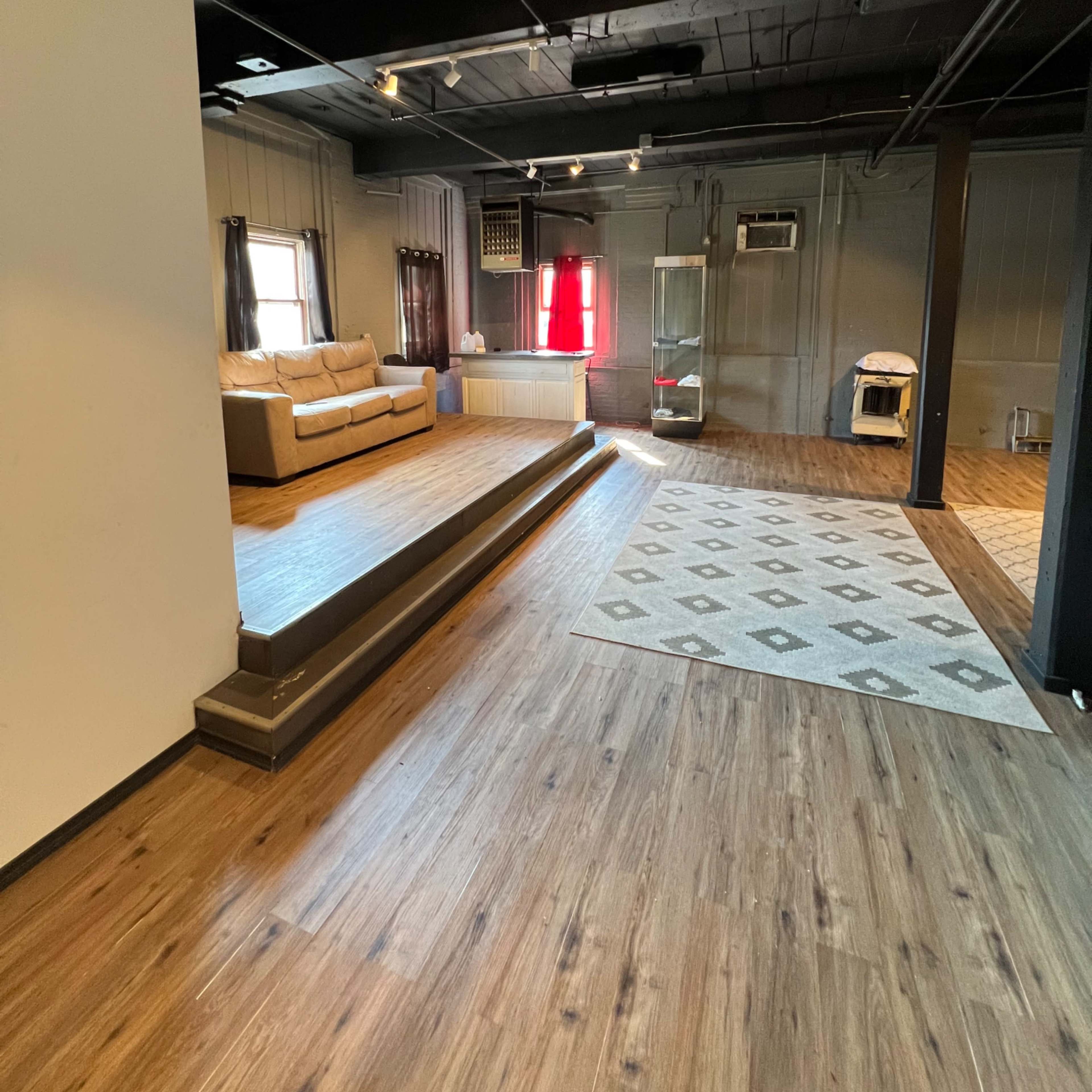 An empty, modern interior space featuring a couch, a small stage area, and a large rug on a wooden floor.