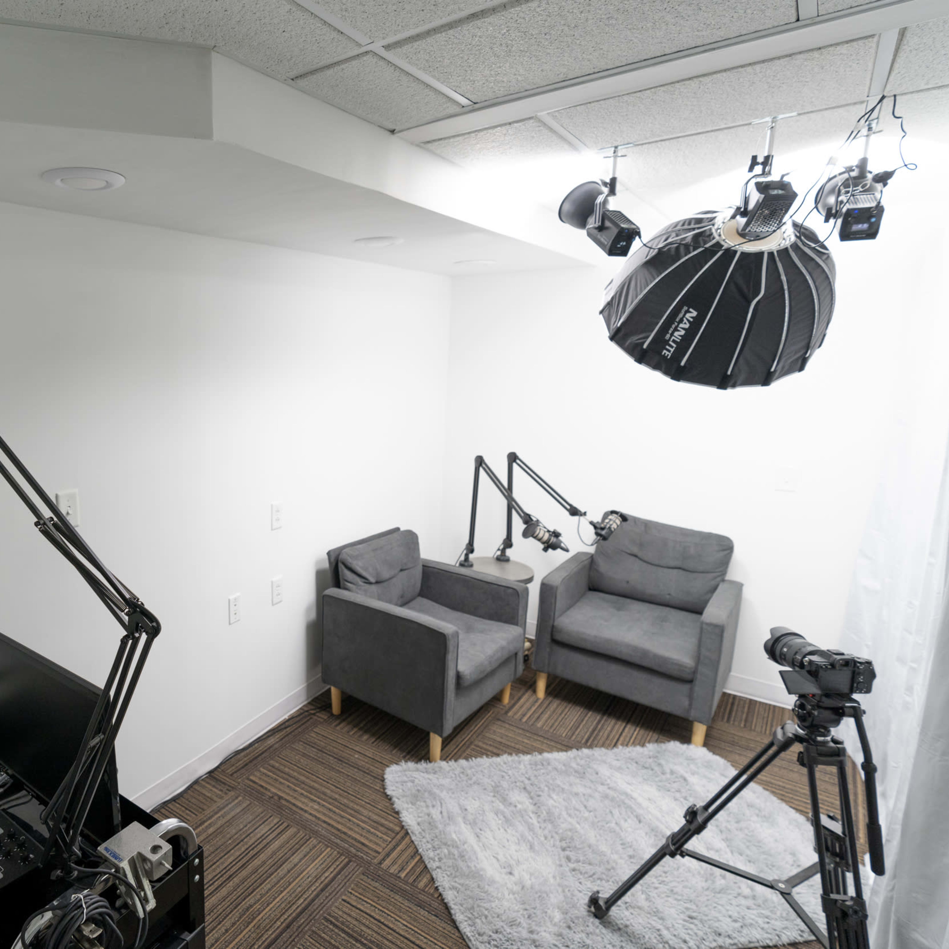 A small recording studio featuring two gray armchairs, a camera on a tripod, a microphone setup, and studio lights overhead.