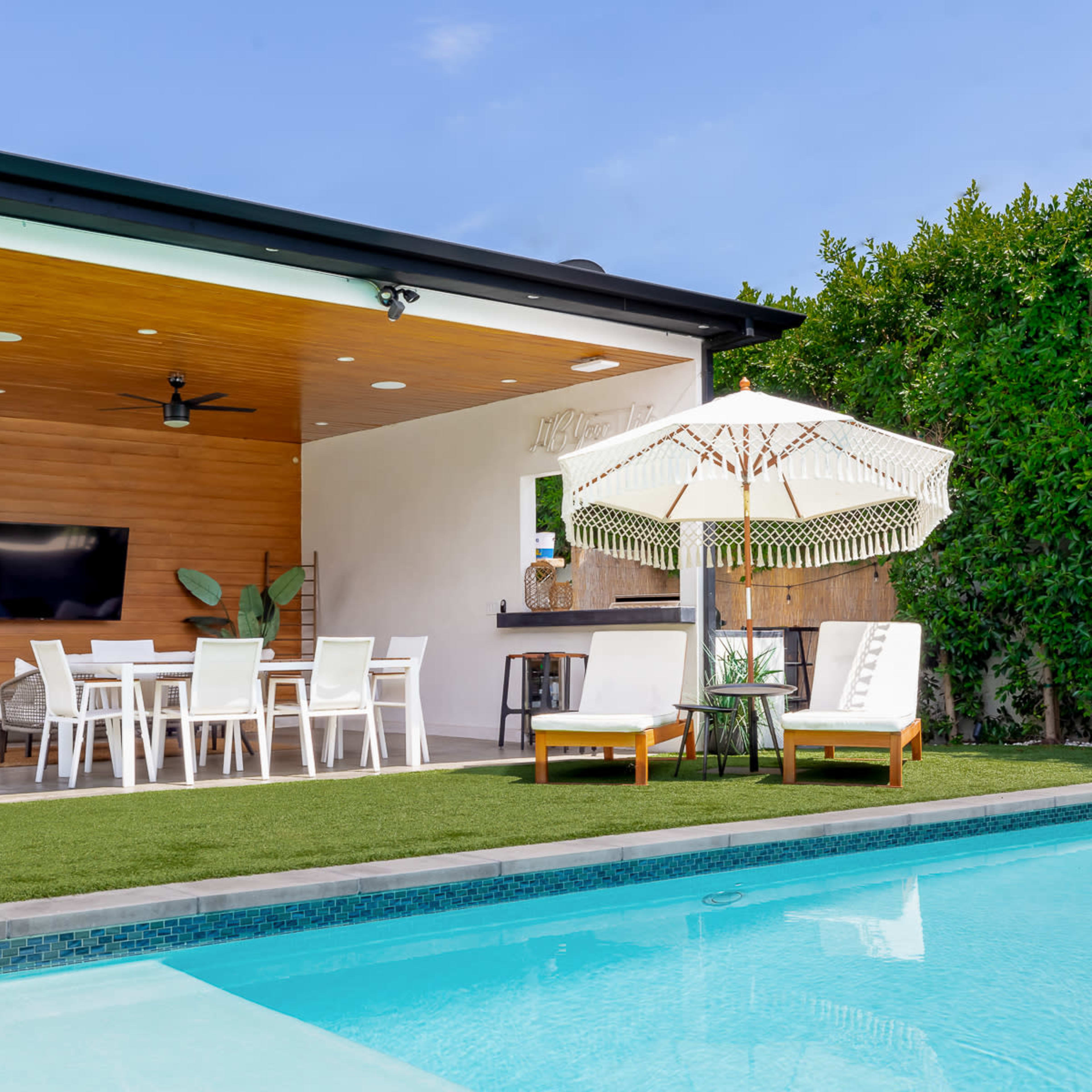 A modern outdoor patio features a dining area, lounge chairs, and a pool surrounded by well-maintained greenery.