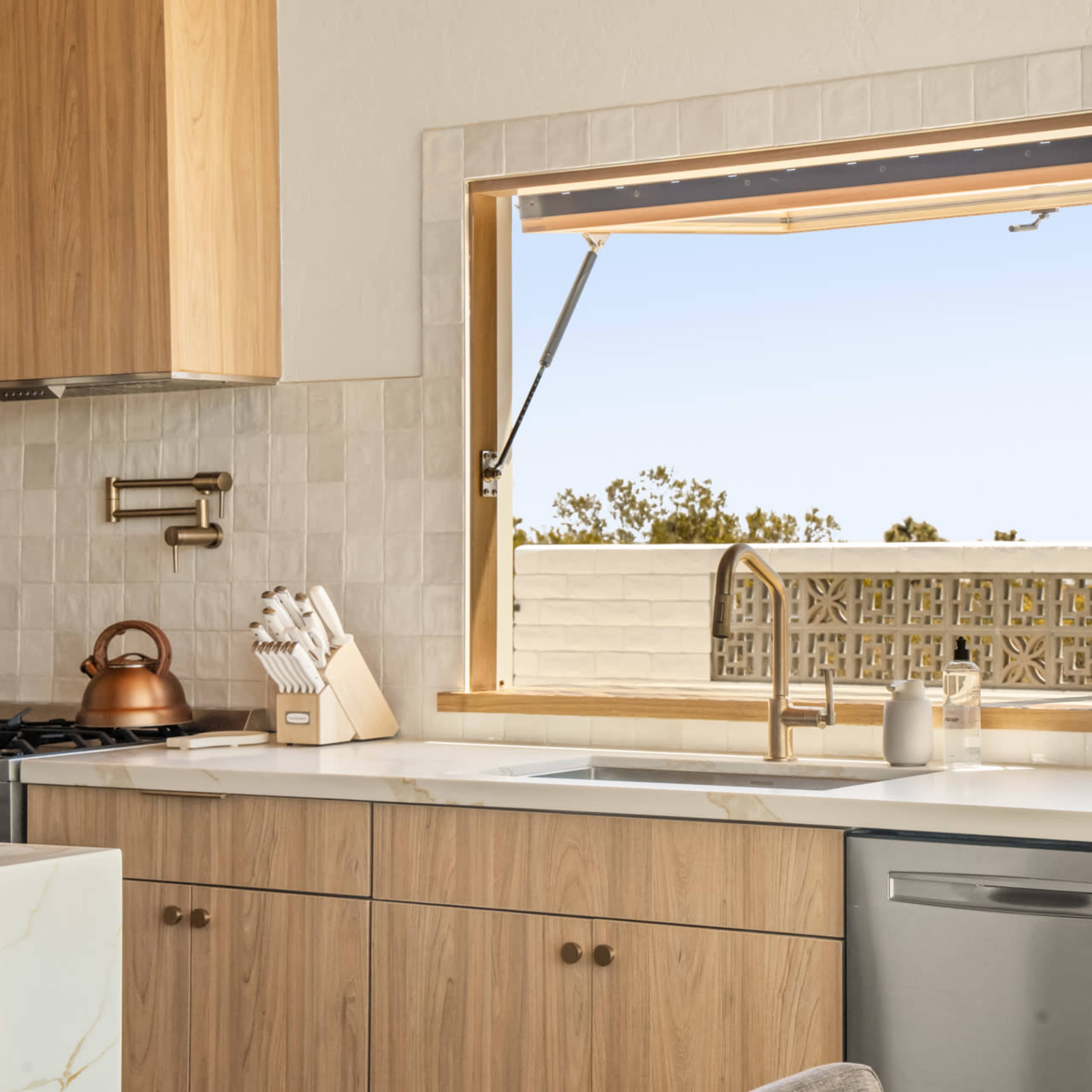 A kitchen features wooden cabinetry, a sink with a sleek faucet, and an open window overlooking a view outside.
